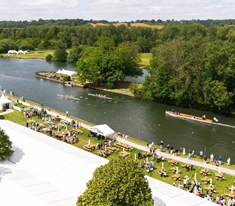 A waterfront park with people near tents and picnic areas, and boats on a river surrounded by trees.