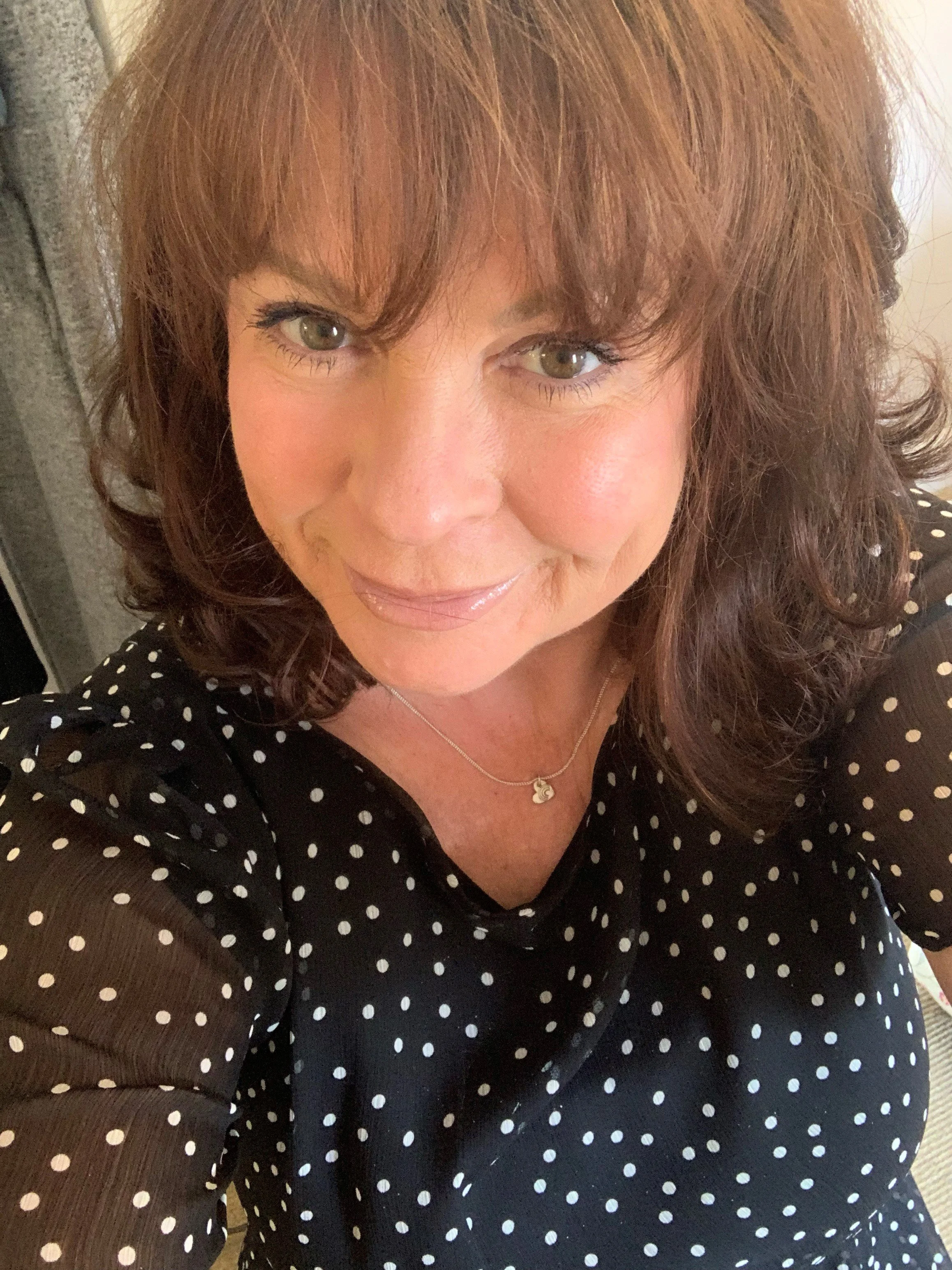 A woman with light skin, brown hair, and green eyes wearing a black dress with white polka dots, sitting indoors near a wall or door, smiling slightly, with a heart-shaped pendant necklace.