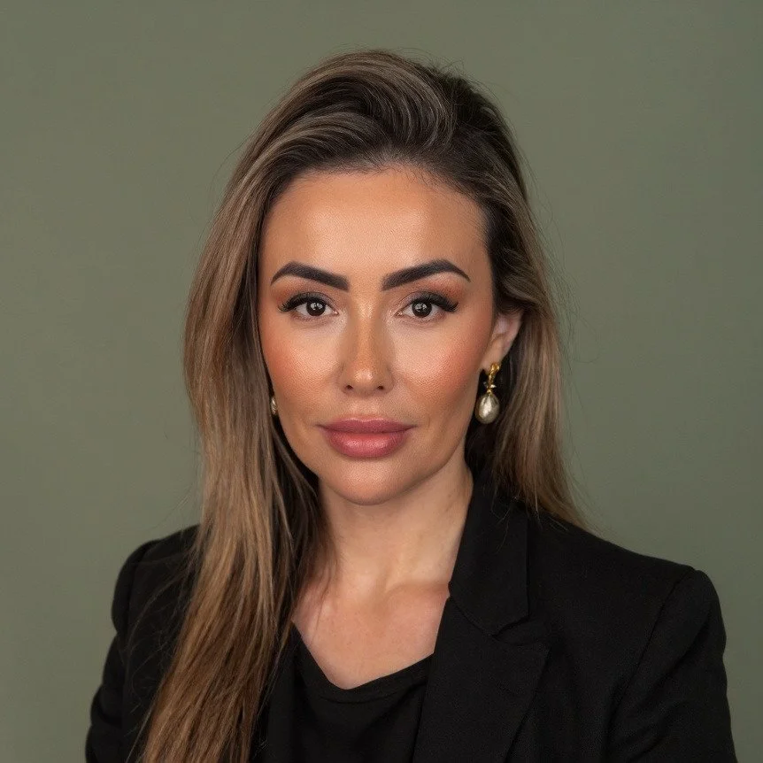 A woman with long brown hair, wearing makeup, gold earrings with pearls, and a black blazer against a plain green background.