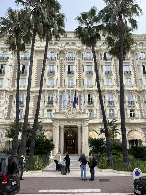 Front view of the Carlton Hotel with a stately, cream-colored façade, palm trees, and people walking and standing in front.