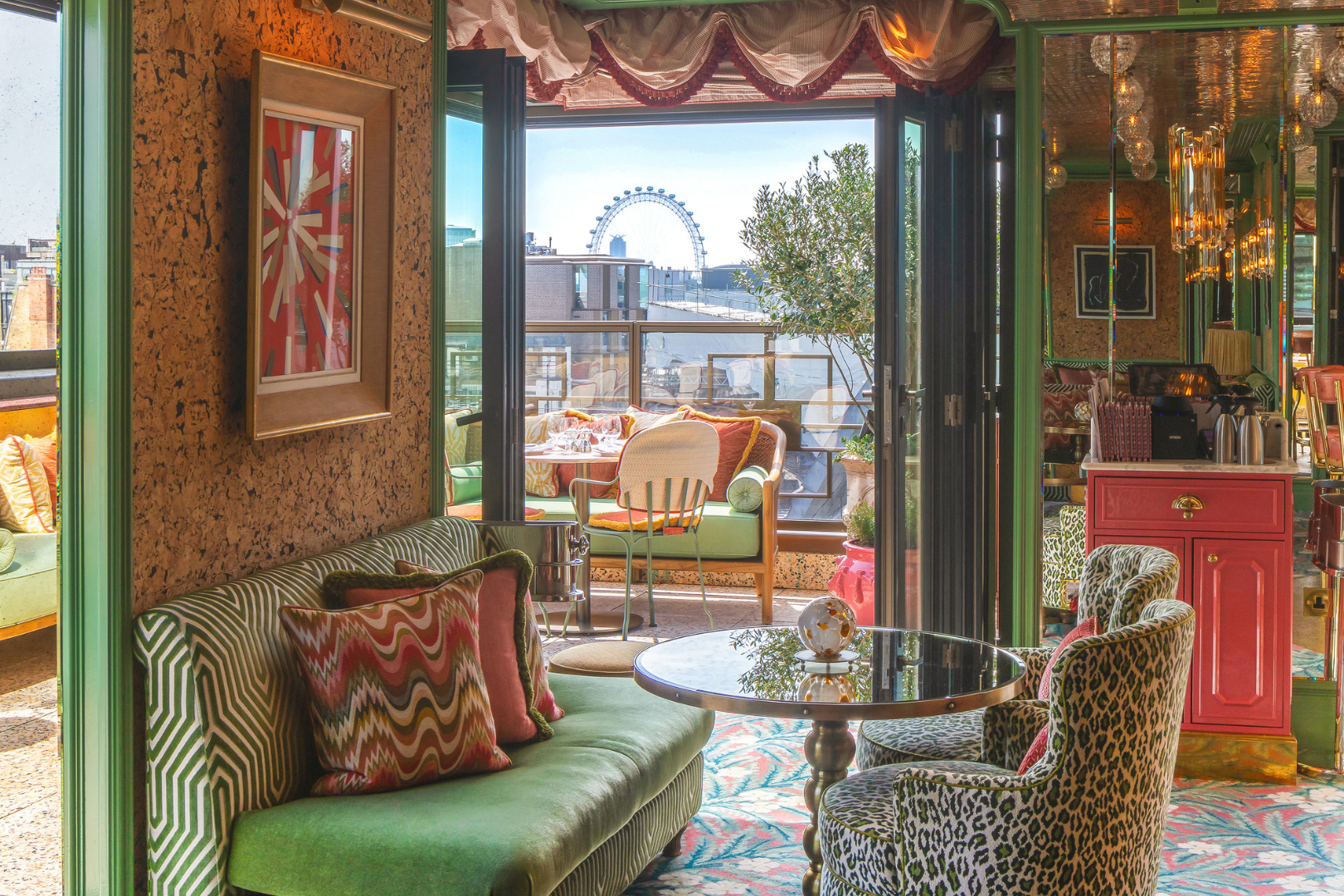 Colorful restaurant or cafe interior with a patio area visible through open sliding doors, with a distant Ferris wheel in the background. The space features eclectic furniture, patterned chairs, a green sofa, pillows, a small pink bar cabinet, and decorative lighting.