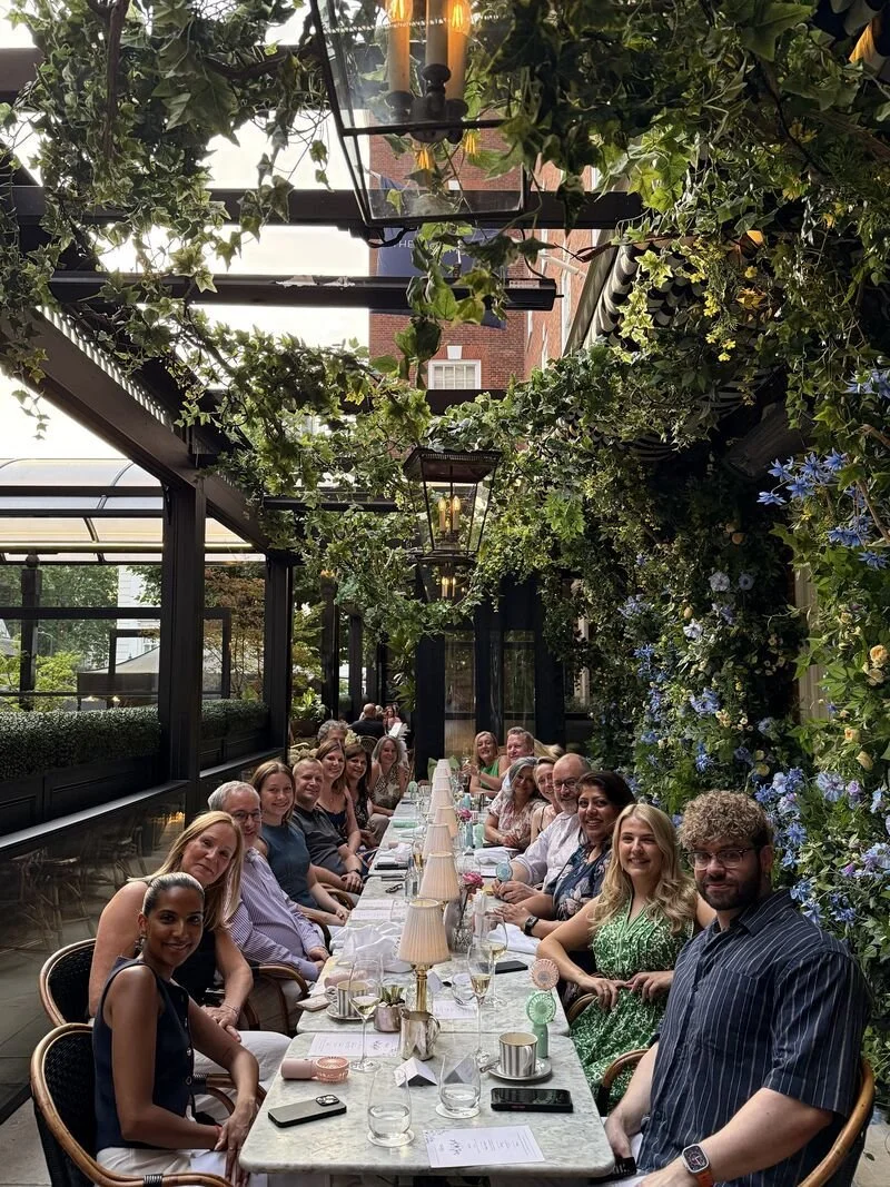 Group of people sitting at a long banquet table decorated with lamps and surrounded by lush green and blue floral arrangements under a covered outdoor patio.