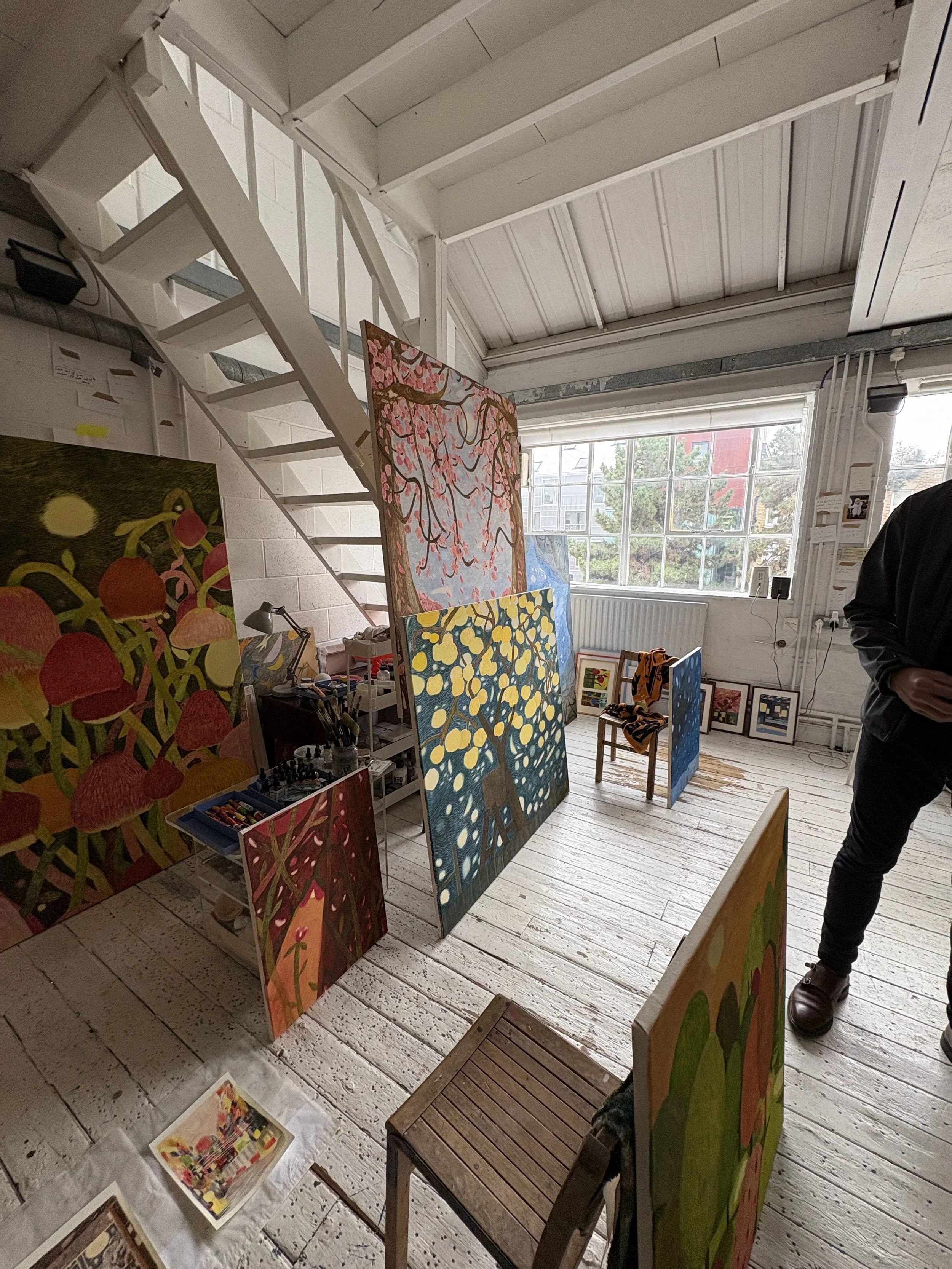 An art gallery with colorful paintings displayed on the floor and leaning against the walls, with large windows allowing natural light to illuminate the room.