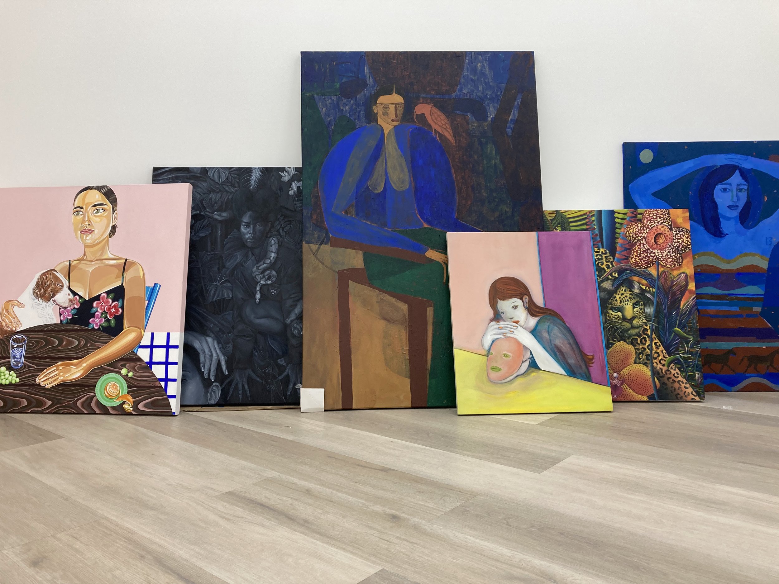 Collection of colorful paintings, including portraits and abstract art, displayed against a white wall. The paintings feature female figures, animals, and vivid patterns.