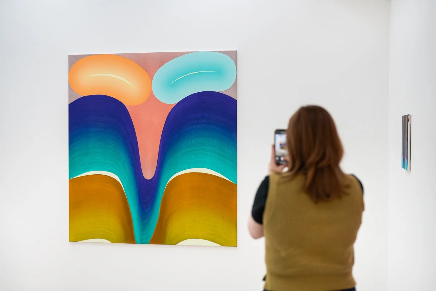 A woman taking a photo of an abstract colorful painting at an art gallery with white walls.