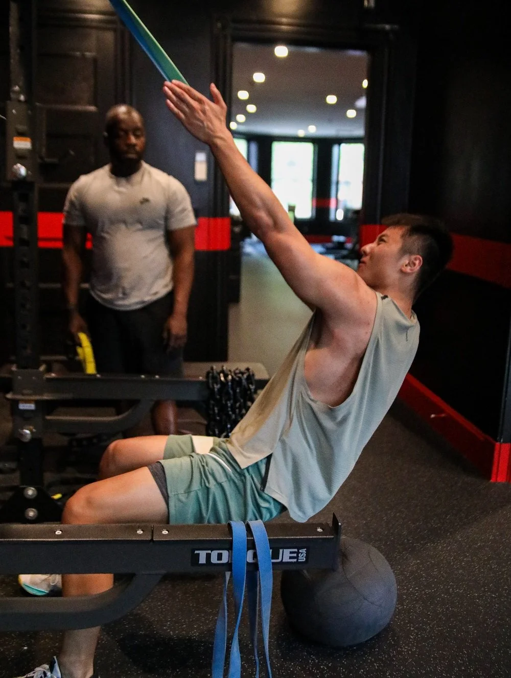 A person exercising with a resistance band on a bench, while another person observes, in a gym setting.