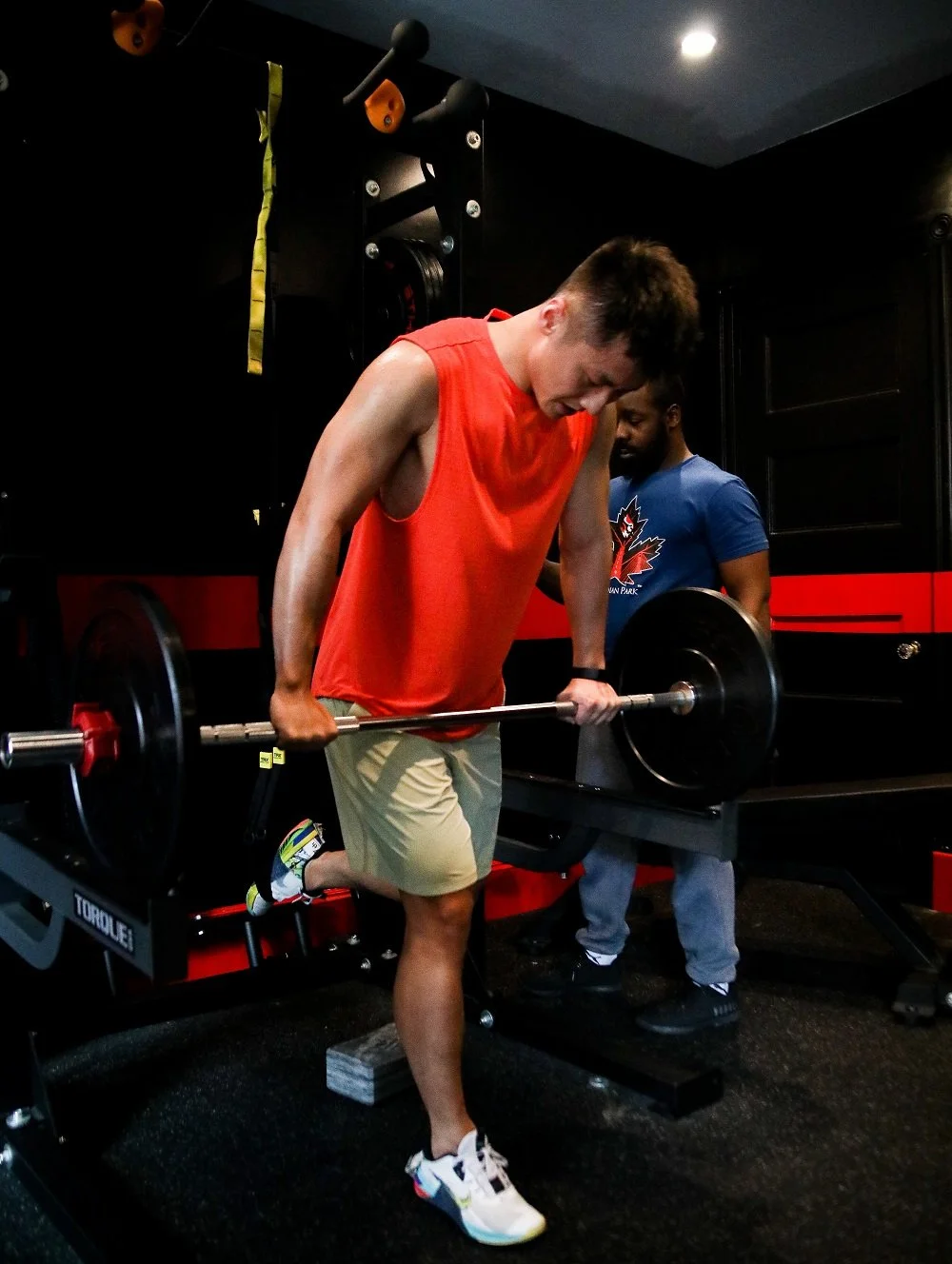 Two people at a gym, one lifting a barbell while the other supervises. The lifter is wearing a red sleeveless shirt and shorts, and the supervisor is in a blue shirt. The setting includes gym equipment and weights.