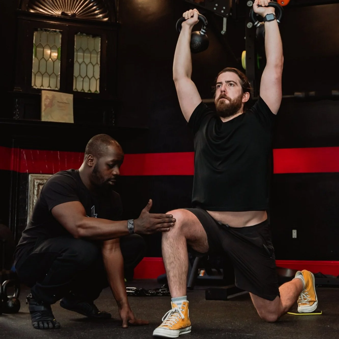 A trainer assists a man performing a kneeling kettlebell exercise in a gym.