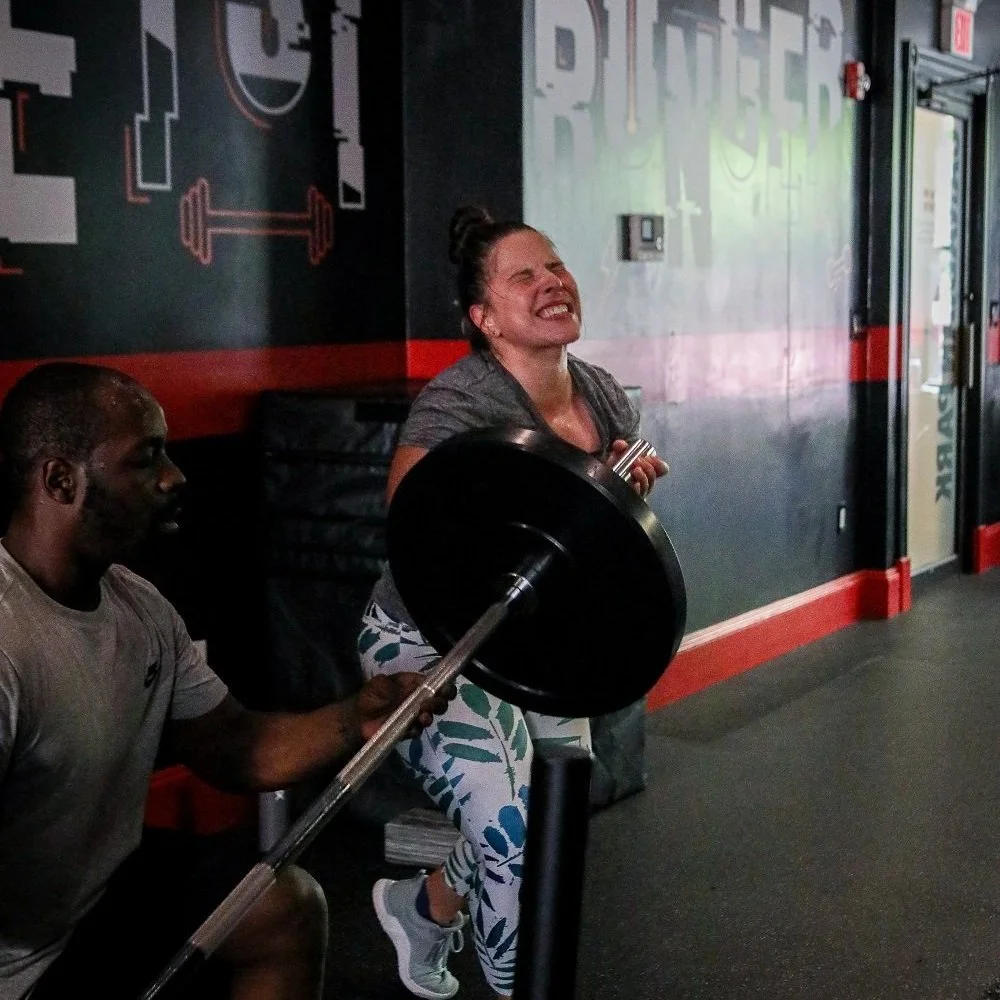 Woman lifting weights with a barbell in a gym, assisted by a spotter. She is exerting effort, wearing floral leggings and a gray top. Gym wall with motivational words in the background.