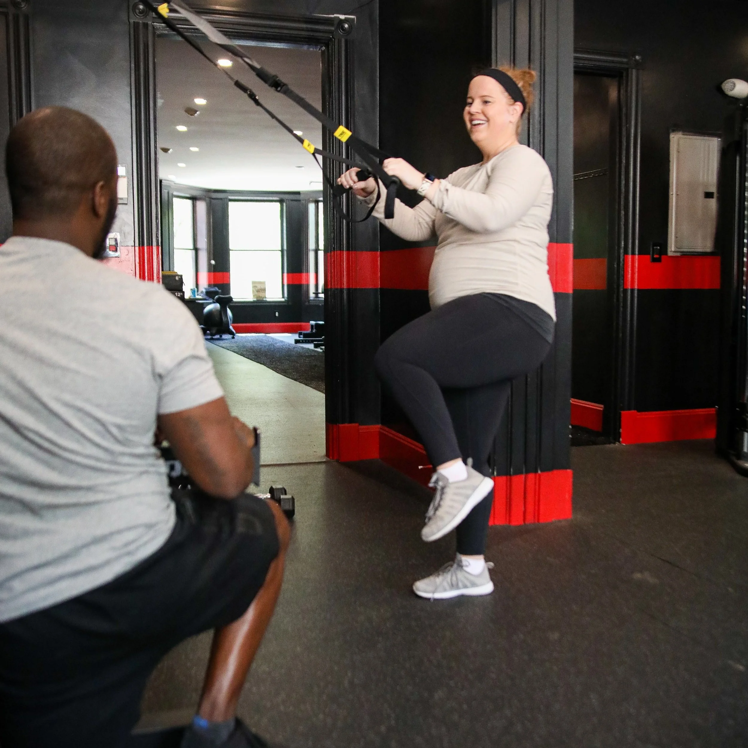 A pregnant woman exercising with resistance bands under the guidance of a fitness trainer in a gym.