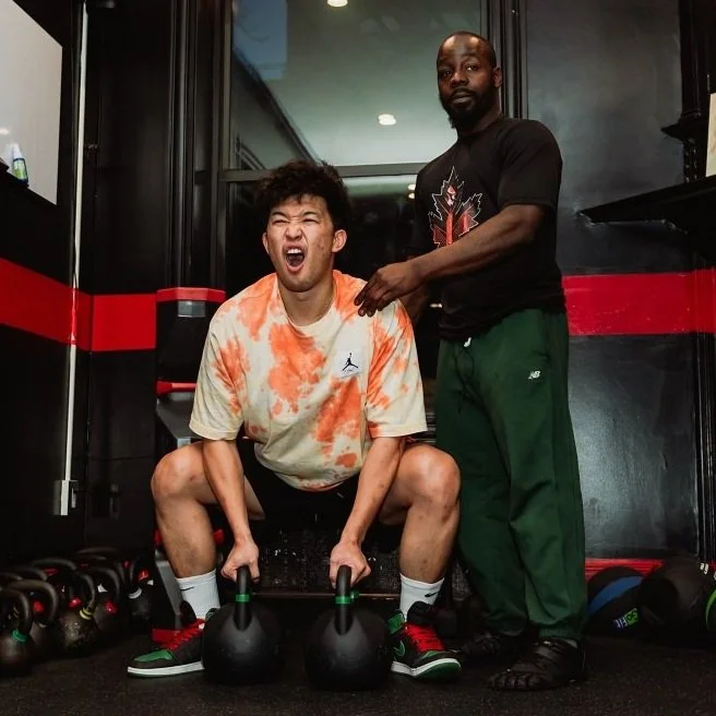 Two men in a gym, one lifting kettlebells with assistance.