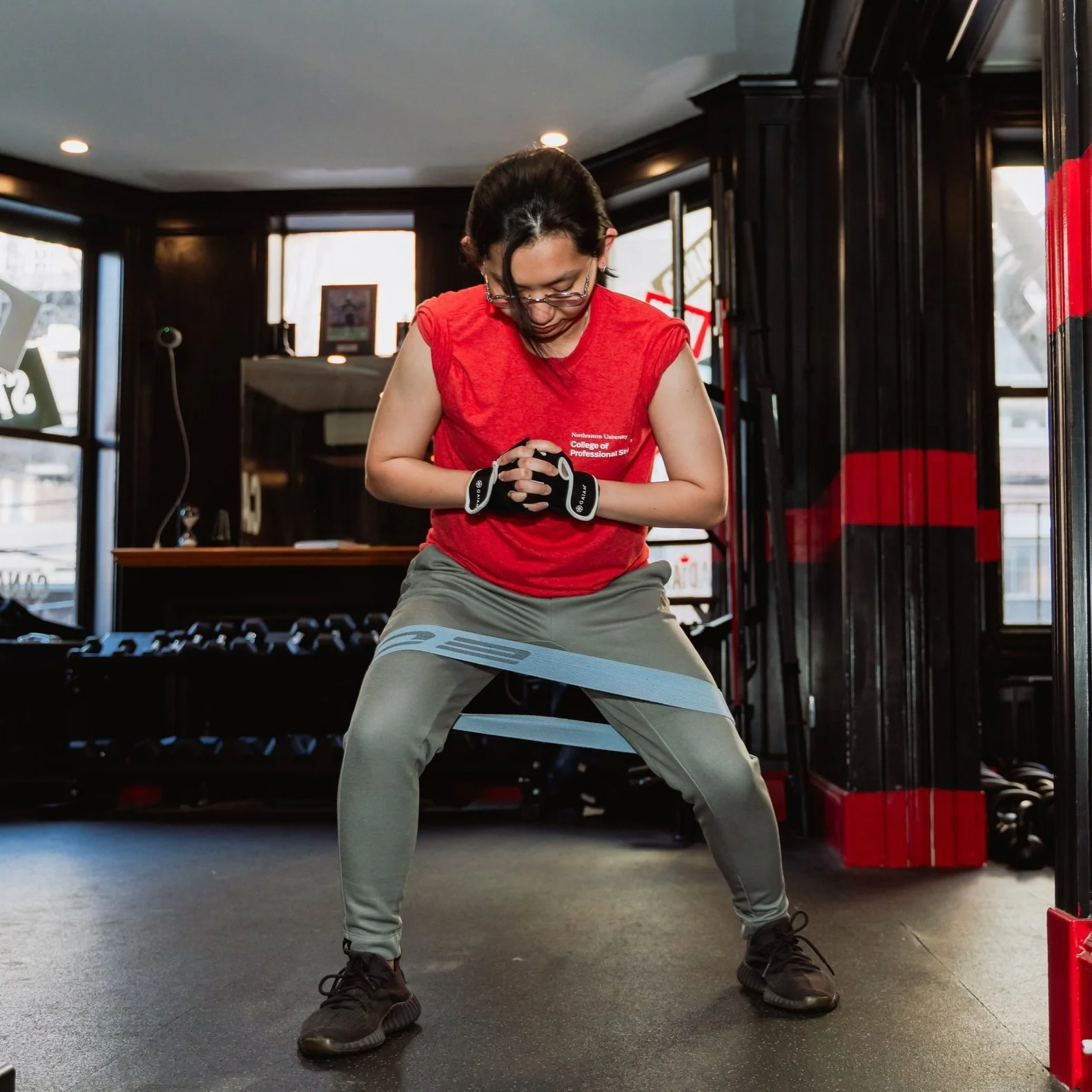 A woman in a red sleeveless shirt and gray pants exercising with a resistance band around her thighs in a gym.