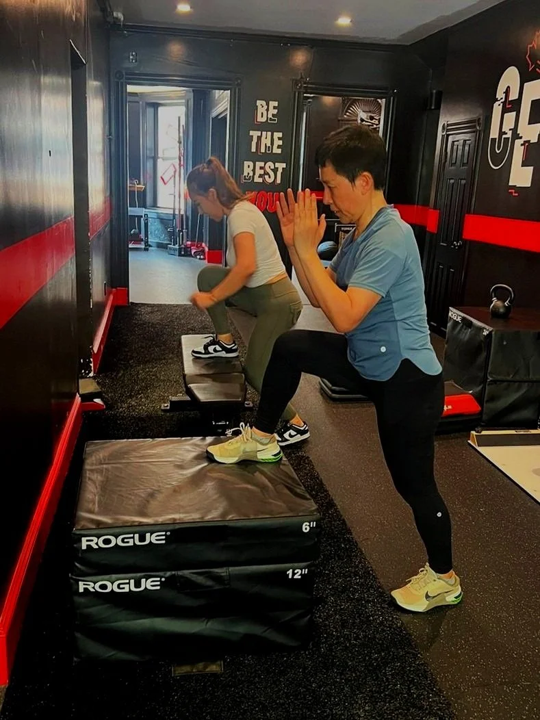 A woman and a man are exercising at a gym, performing step-up exercises on plyometric boxes labeled 6 inches and 12 inches. The woman is stepping onto the smaller box while the man stands next to it with hands together in a prayer position. The gym has black walls with red accents, motivational signs, and workout equipment in the background.