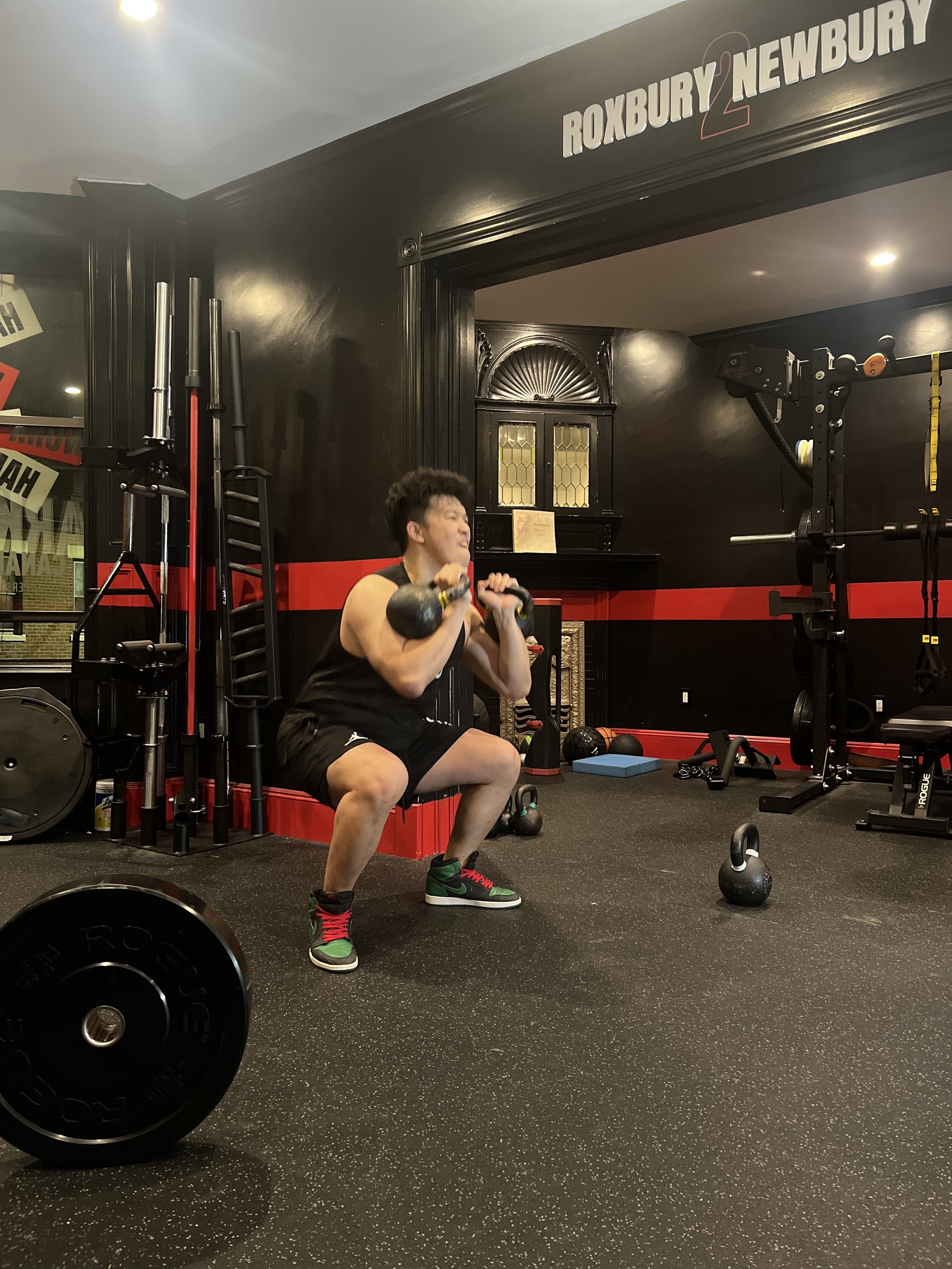A person performing a squat exercise with a kettlebell in a gym with black and red walls and various gym equipment.