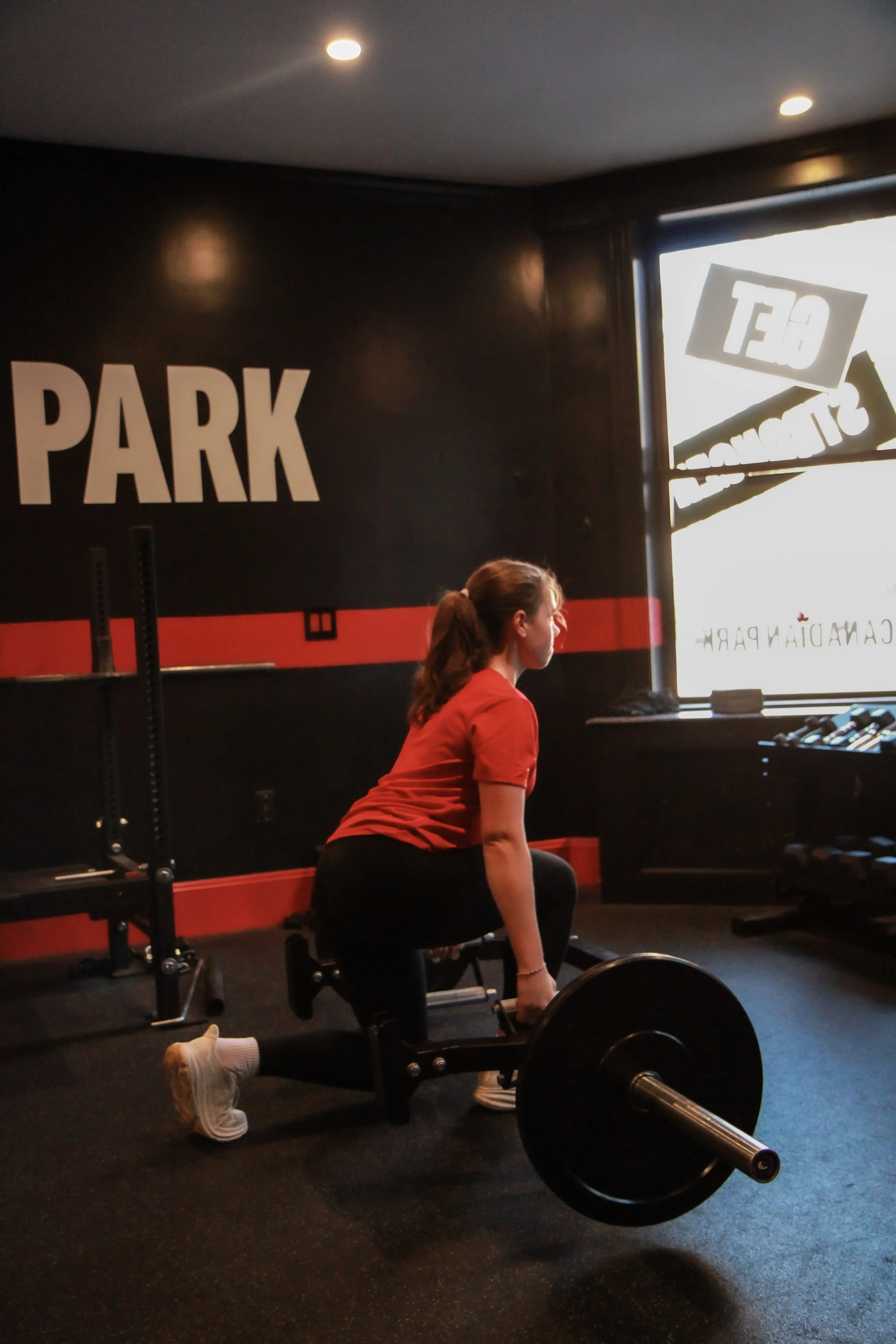 A woman in a red shirt and black leggings is performing a deadlift exercise with a barbell inside a gym, near a wall with the word 'PARK' painted on it.