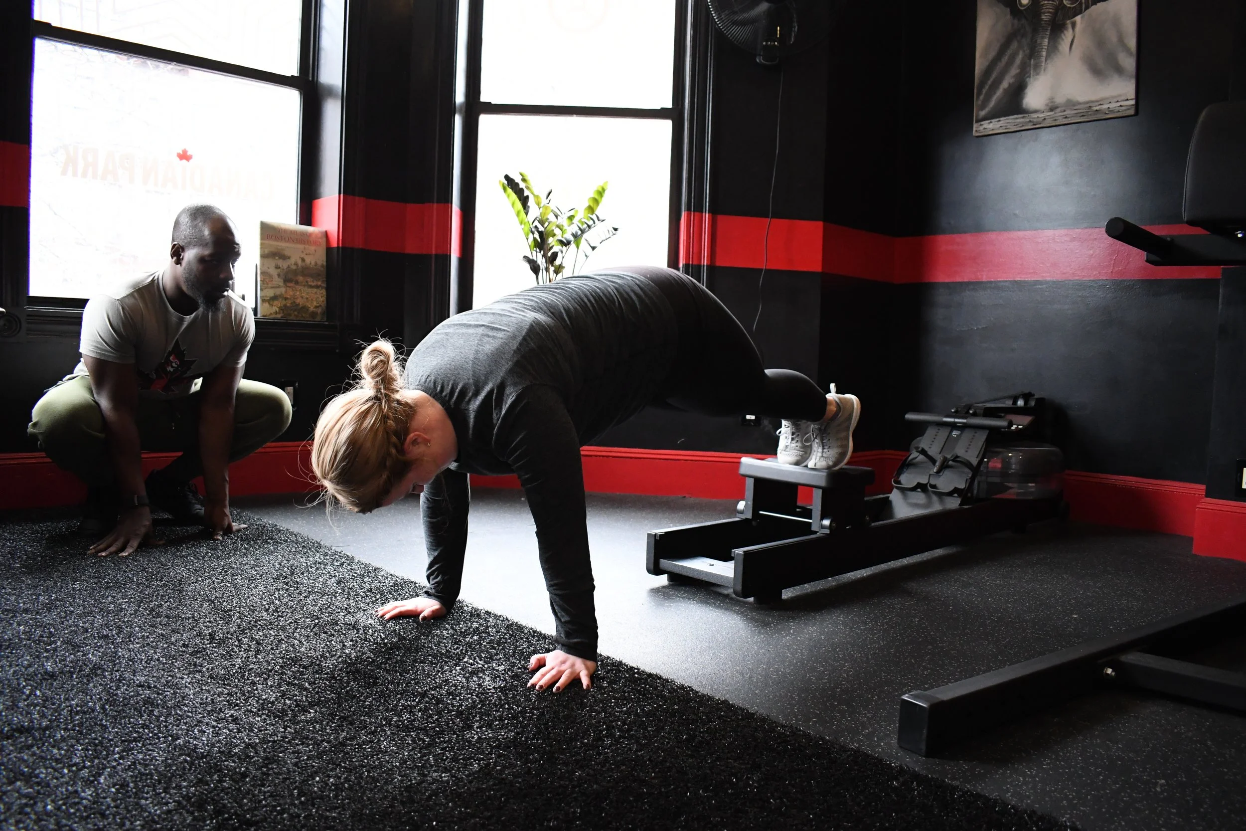 A woman is doing a push-up exercise with her feet elevated on a step in a gym, while a trainer observes her gently from the side.