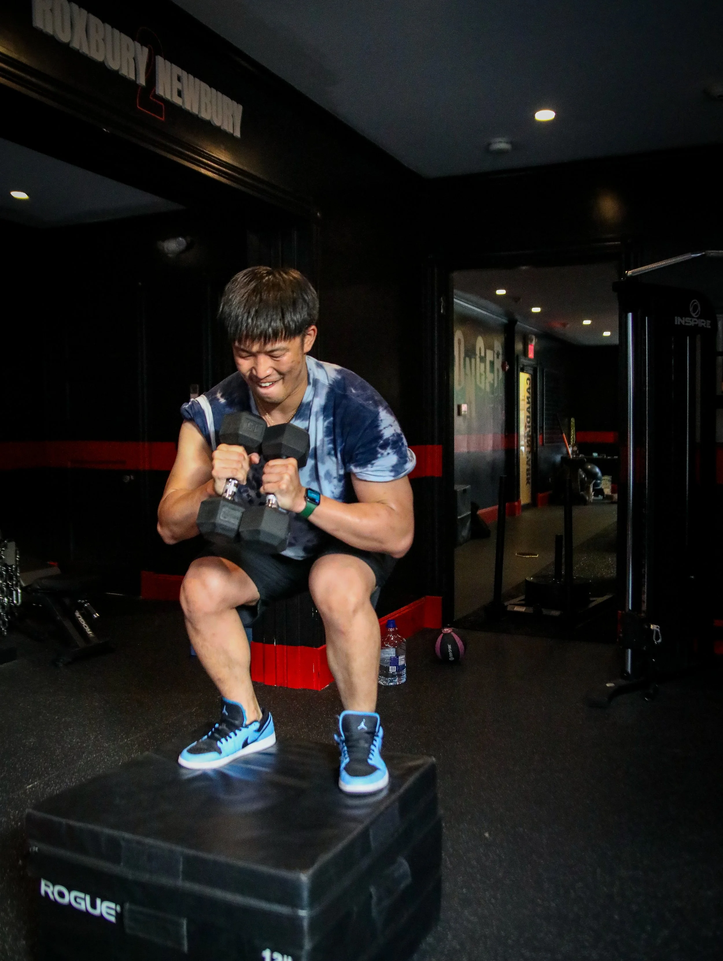 A man in a gym setting sitting on a box, smiling and lifting dumbbells while engaging in a workout.