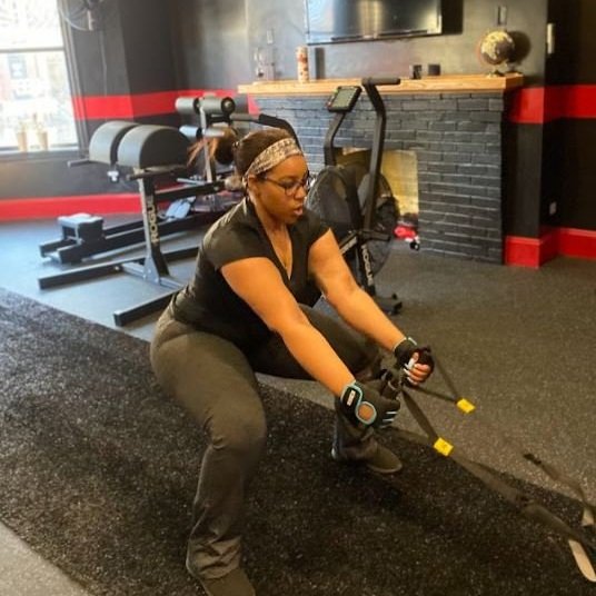 A woman using battle ropes in a gym, crouched down with a focused expression, wearing workout gloves, glasses, and a headband.