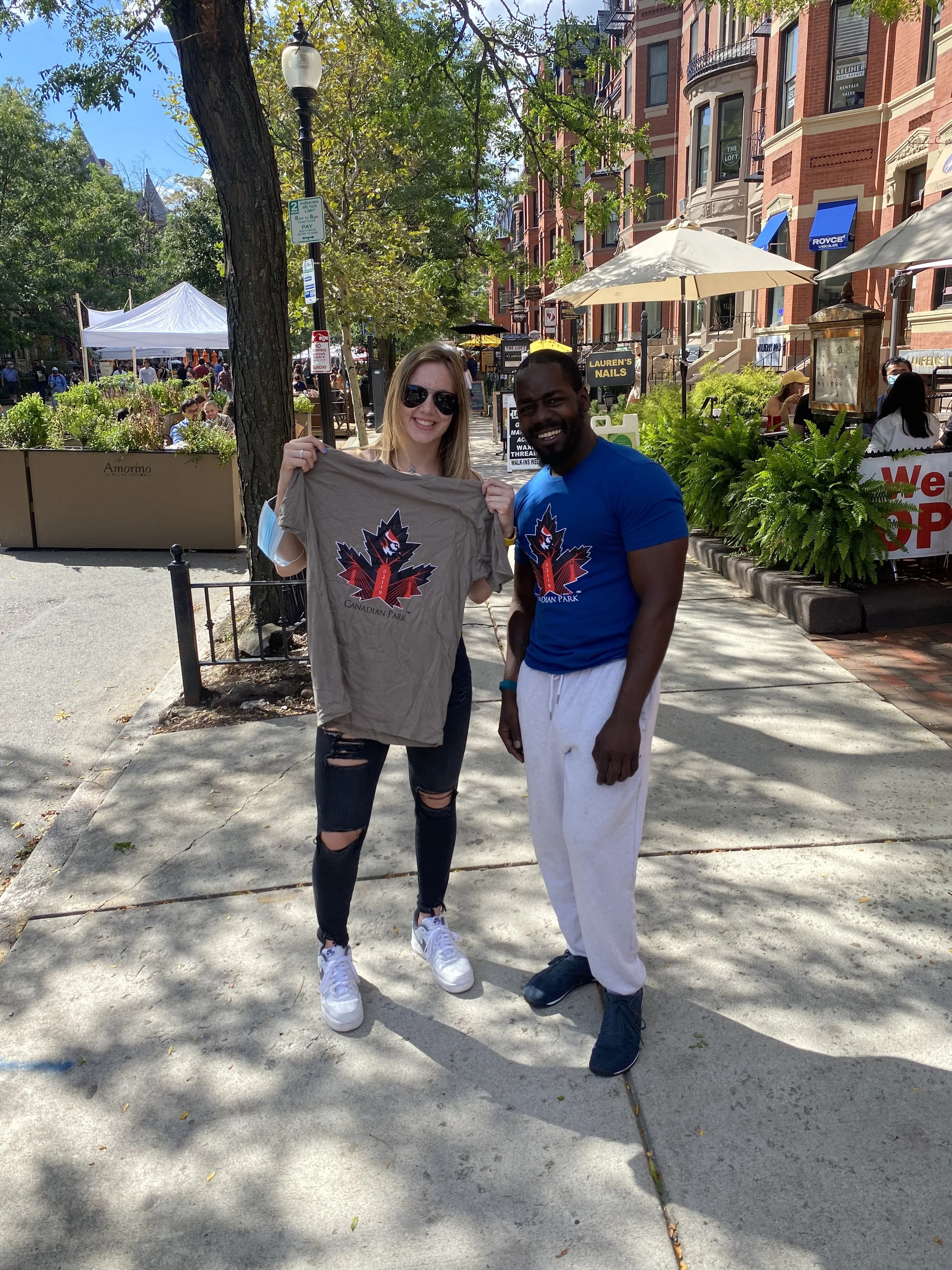 A woman and a man standing on a sidewalk in a city. The woman is holding a gray t-shirt with a logo that says 'Canadian Park' with a red and black maple leaf emblem. The woman is wearing black ripped jeans, a white shirt, and sunglasses. The man is dressed in a blue t-shirt and white sweatpants. They are smiling, and there are outdoor tables and a busy street with trees, buildings, and tents in the background.