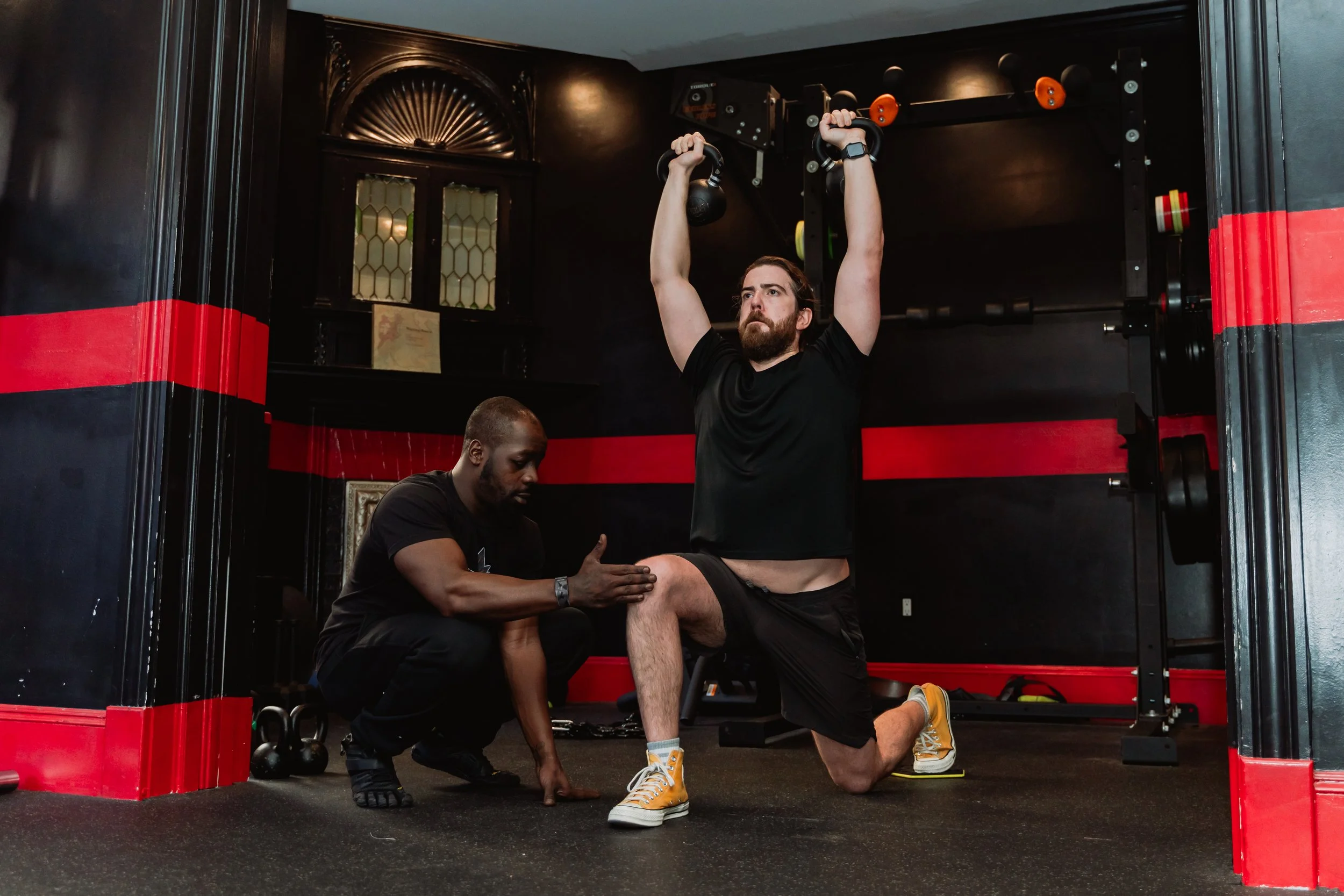 A man is doing a kneeling overhead press with kettlebells supervised by a trainer in a gym with black and red walls.