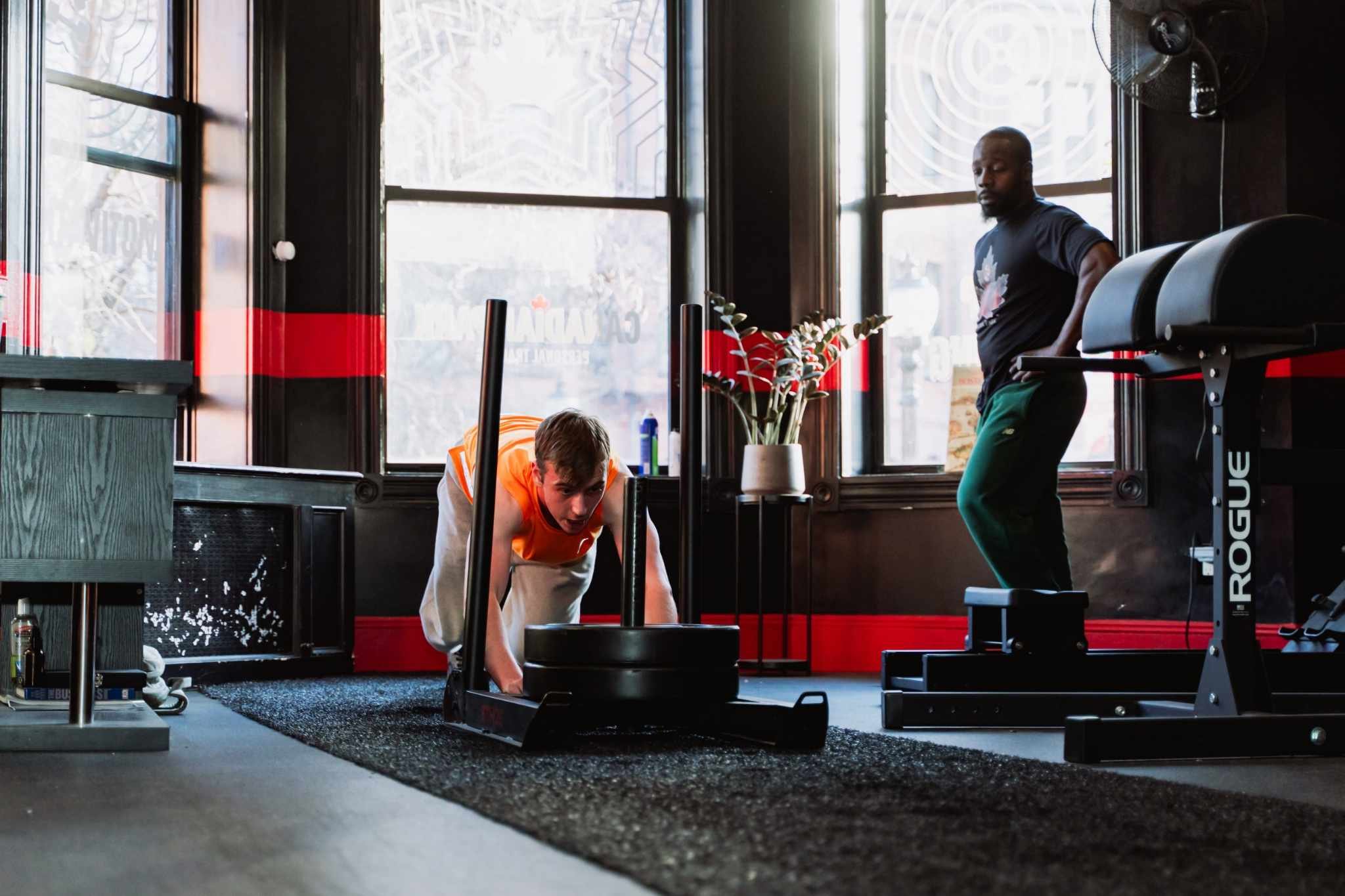 Two people in a gym; one is pushing a sled and the other is standing by gym equipment.
