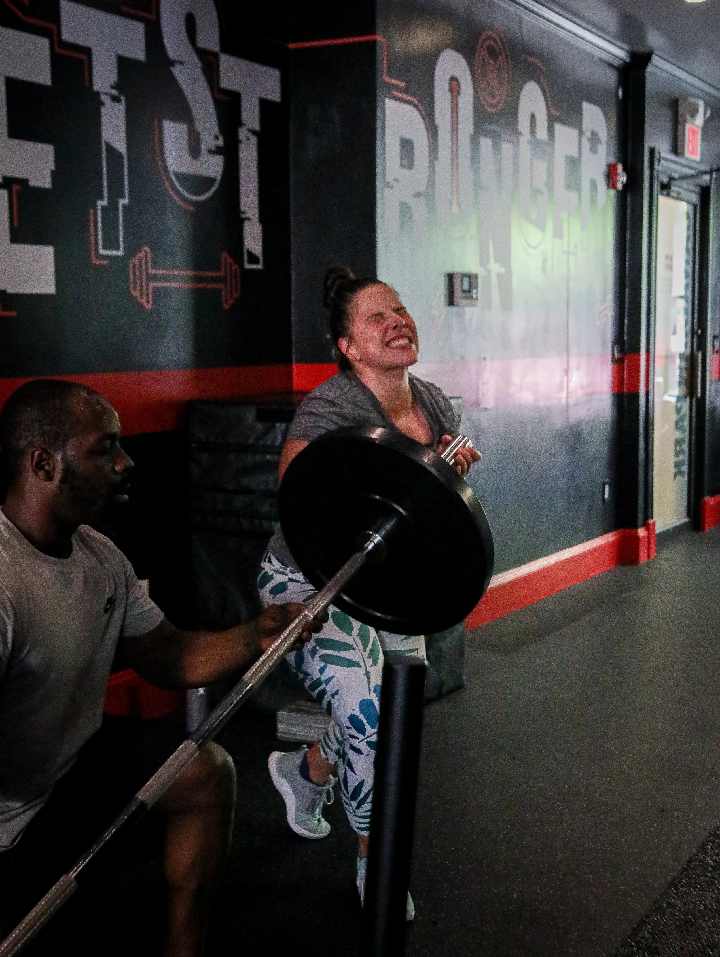 A woman lifting a barbell with weights under the guidance of a trainer at a gym.