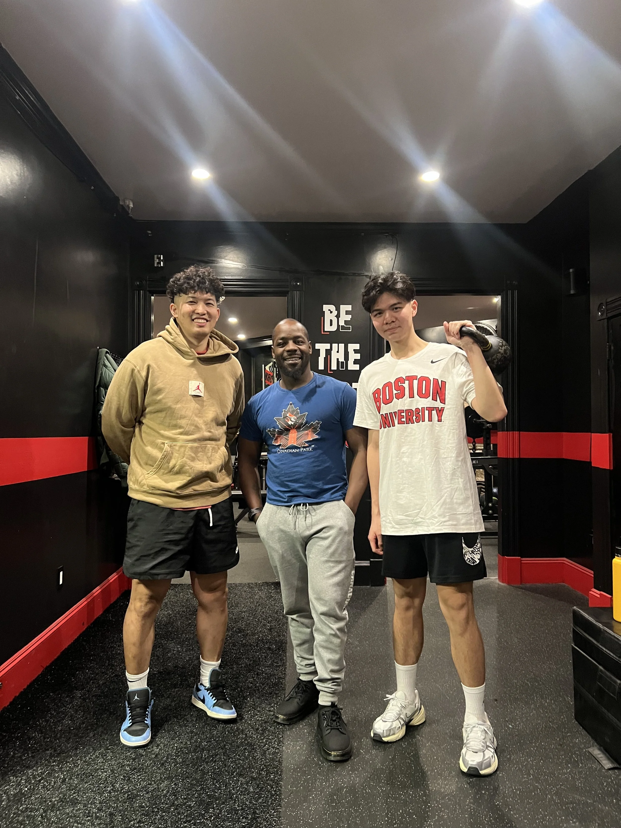 Three men standing in a gym with fitness equipment, smiling and looking at the camera.