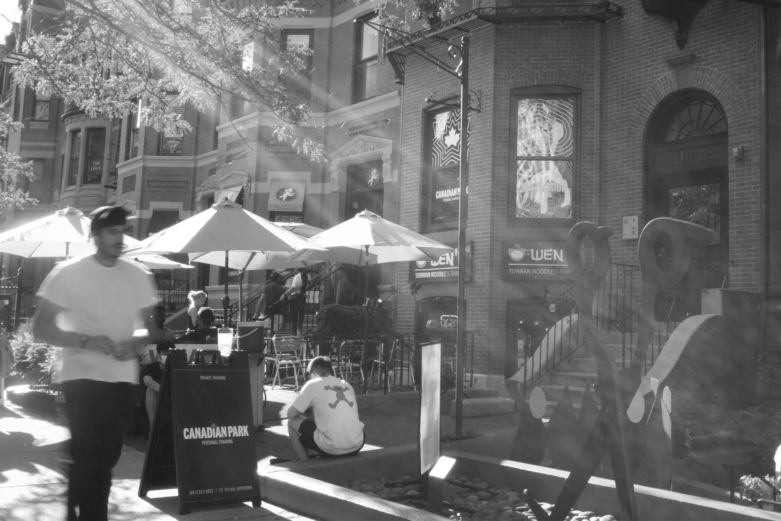 Street scene outside a restaurant with outdoor seating, umbrellas, and people, including a person sitting and others walking, reflected in a large window.