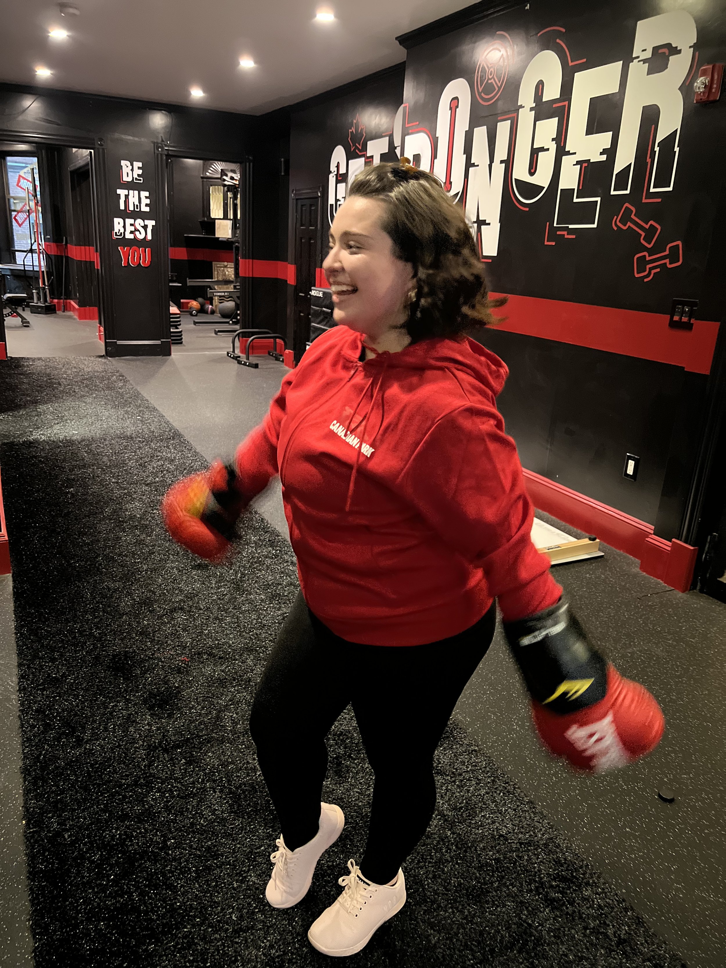 A woman in a red hoodie and black leggings is smiling and holding red boxing gloves inside a gym with black walls and motivational quotes.