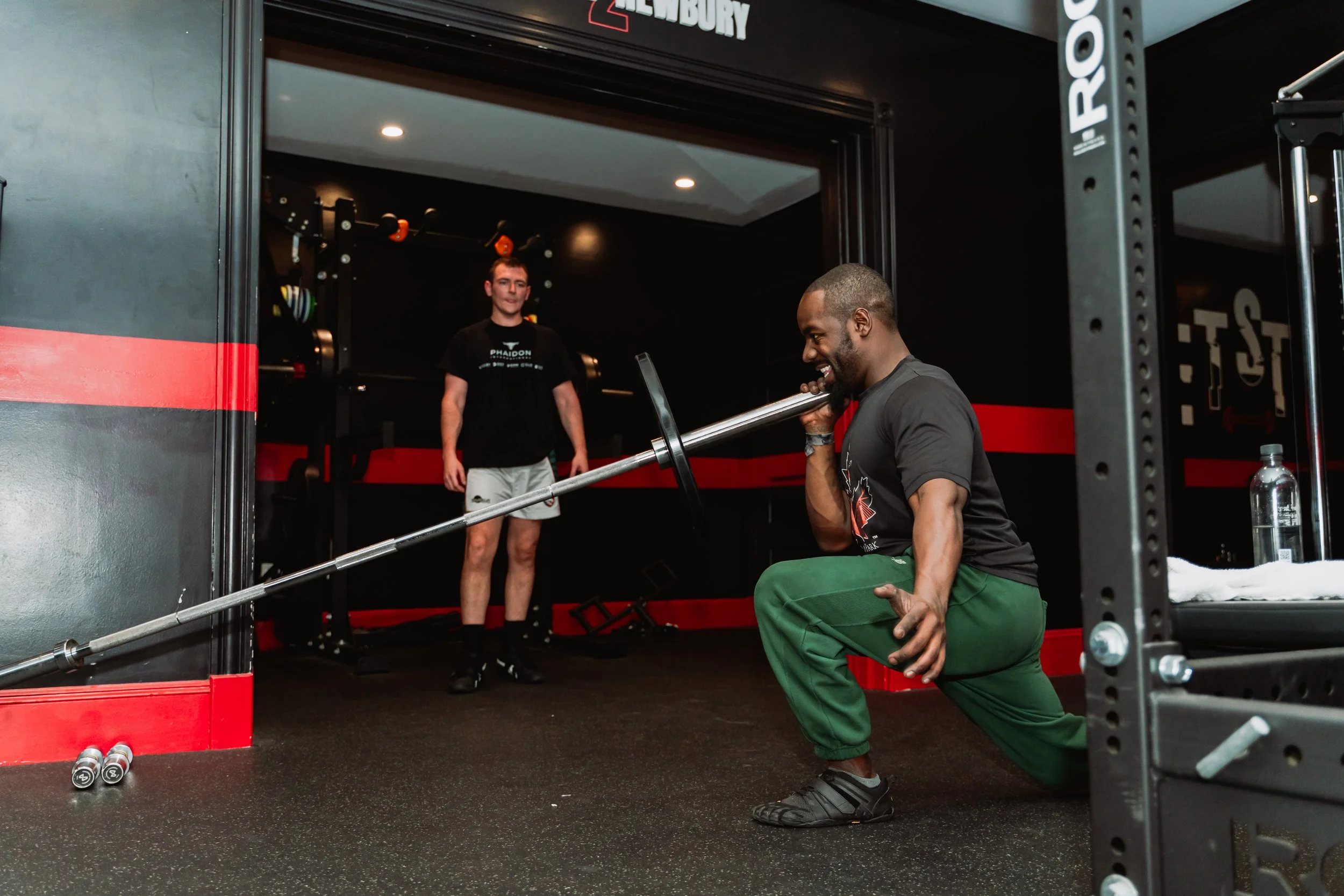 A man is performing a squat exercise with a barbell in a gym while another man observes, standing in the background, near gym equipment, with black walls and red accents.