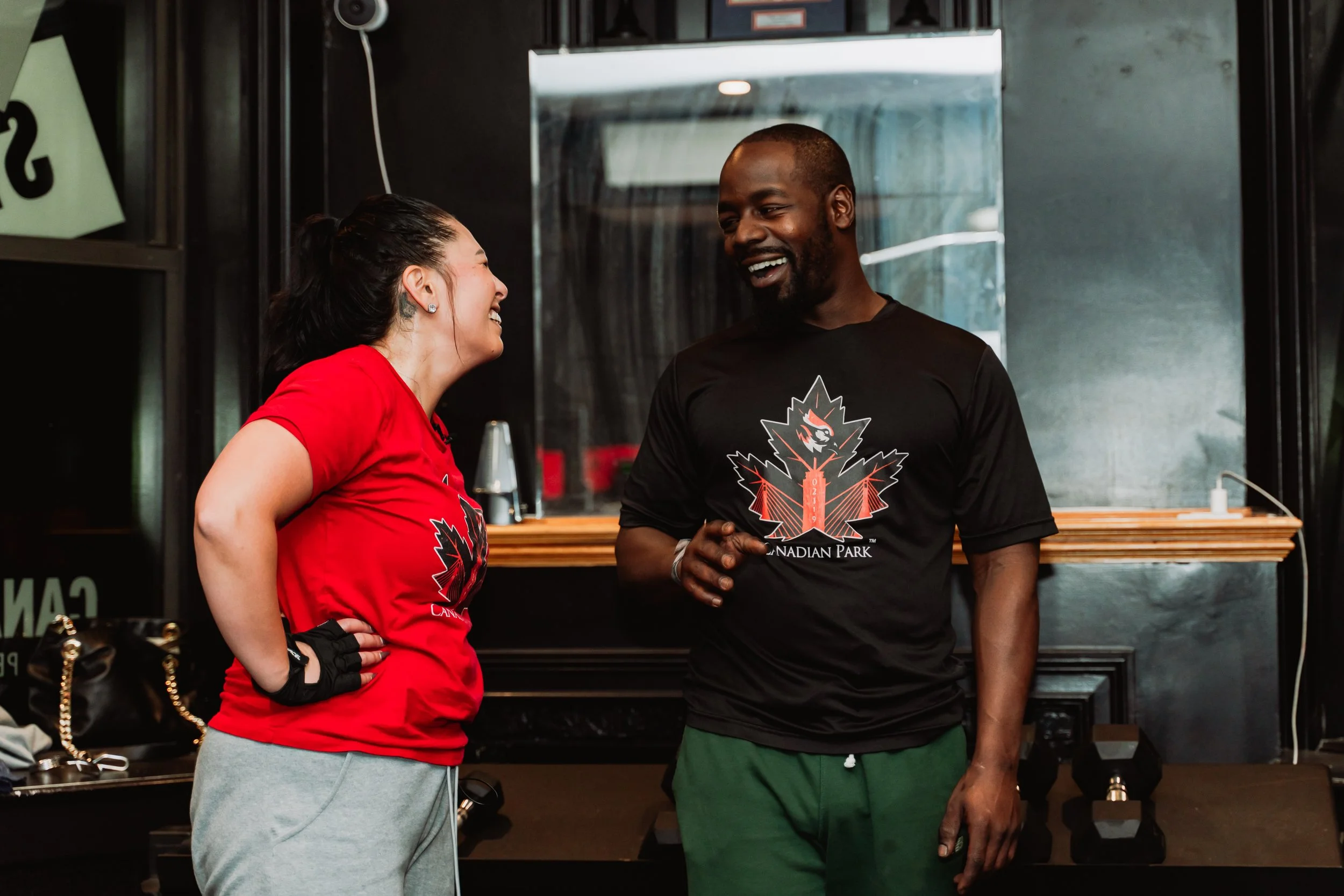 A woman and a man are smiling and talking to each other inside a building. The woman is wearing a red shirt and gray pants and has tattoos on her neck and earrings. The man is wearing a black Canadian Park t-shirt and green shorts. There are black walls and a mirror in the background.