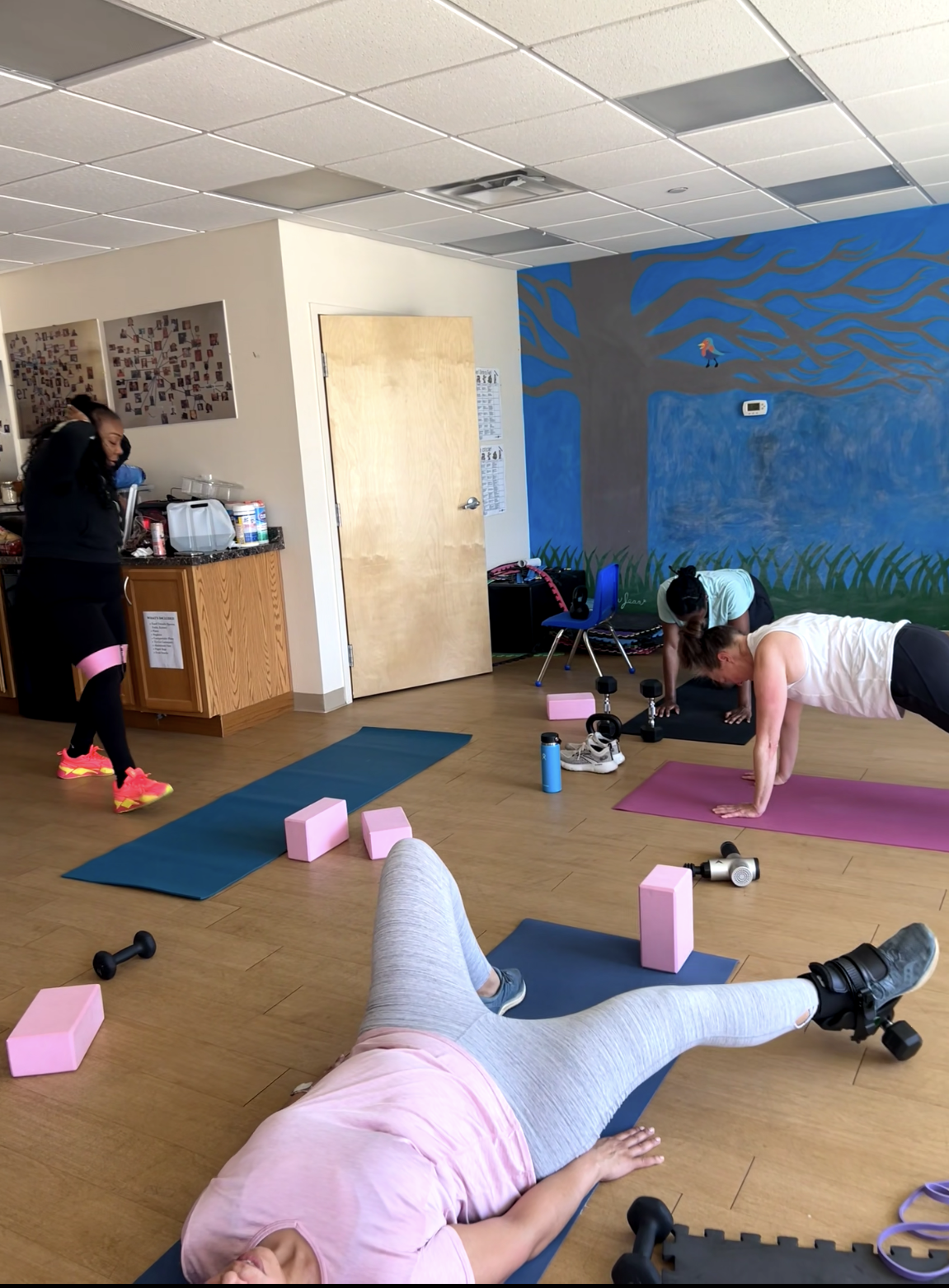 A group exercise class in a room with a colorful mural of a tree on the wall. Participants are using yoga mats, pink foam blocks, and small dumbbells for strength and flexibility workouts. One person is lying on a yoga mat, while others are in plank positions or moving around.