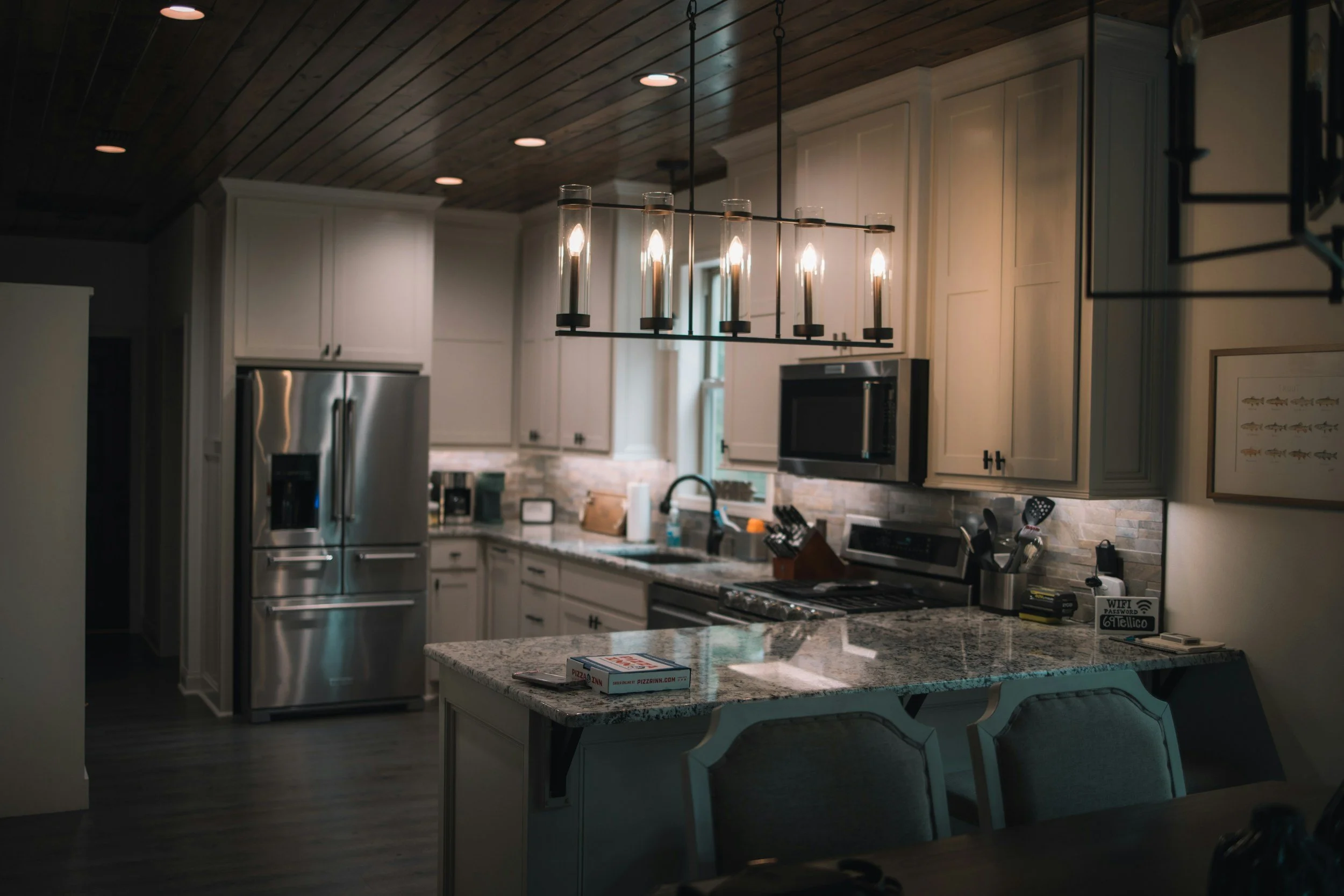 Modern kitchen with white cabinets, stainless steel refrigerator, granite countertops, and a wooden ceiling. A large black and glass chandelier hangs above the island, which has a pizza box on it. The kitchen is well-lit with natural and artificial light.