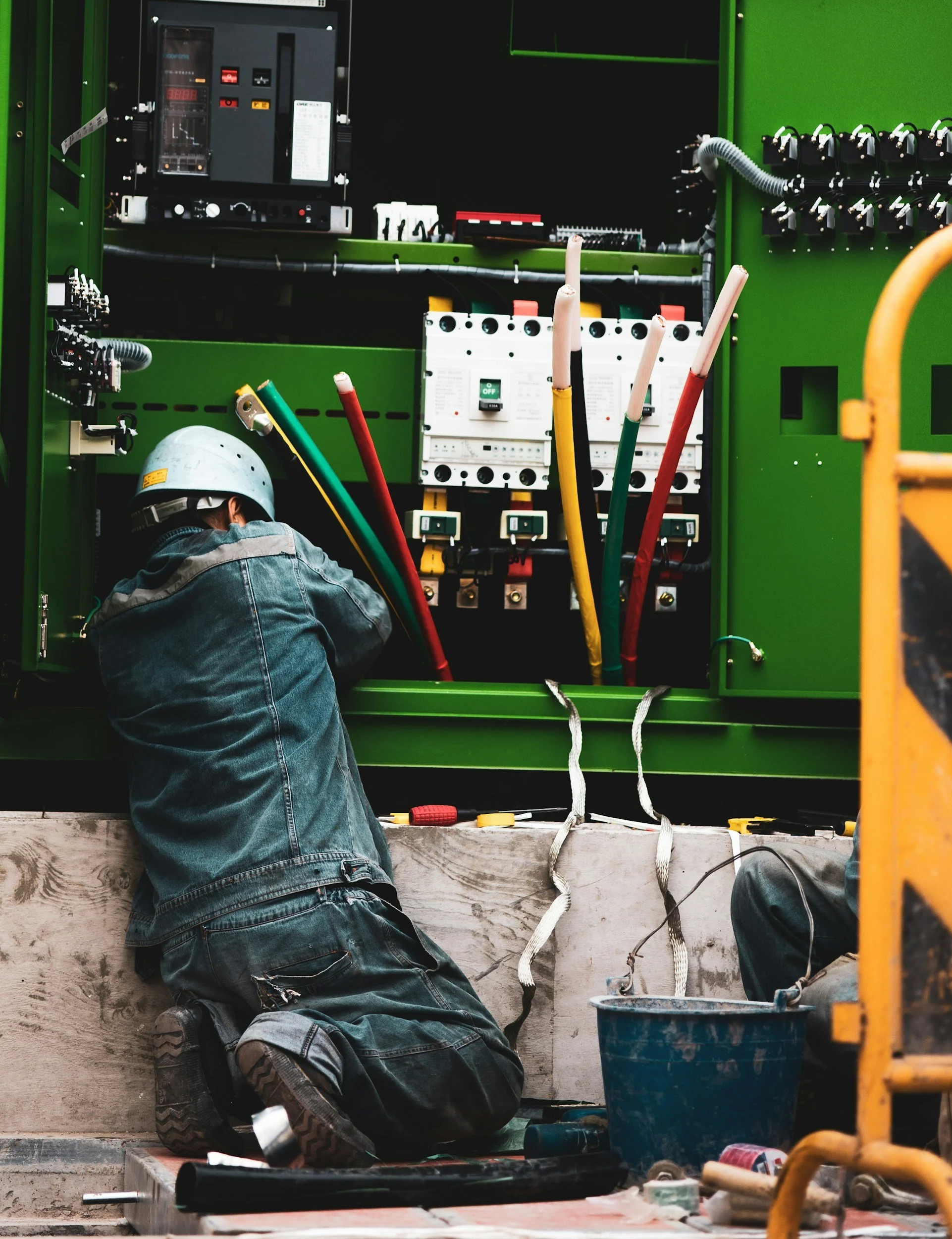 Worker in a hard hat and denim jeans repairing electrical equipment inside a green electrical cabinet with wires and circuit breakers.