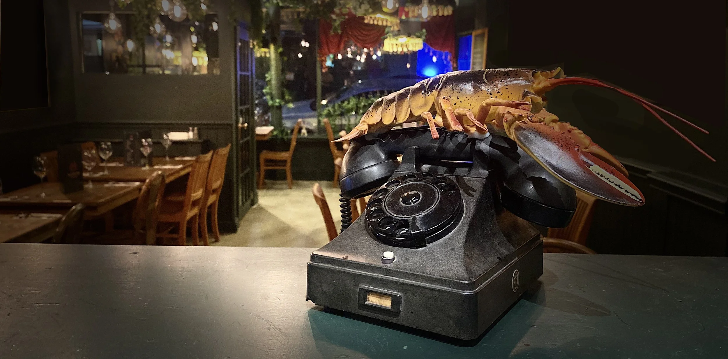 A live lobster with long antennae resting on an old black rotary telephone, placed on a dark surface inside a restaurant with wooden tables and chairs. The background shows large windows with a view of an outdoor area illuminated by string lights and colored decorations.