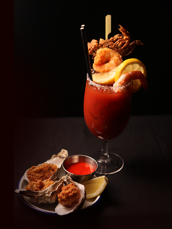 A Bloody Mary cocktail garnished with shrimp, lemon, and fried seafood, served with a side of oysters and a small cup of cocktail sauce.