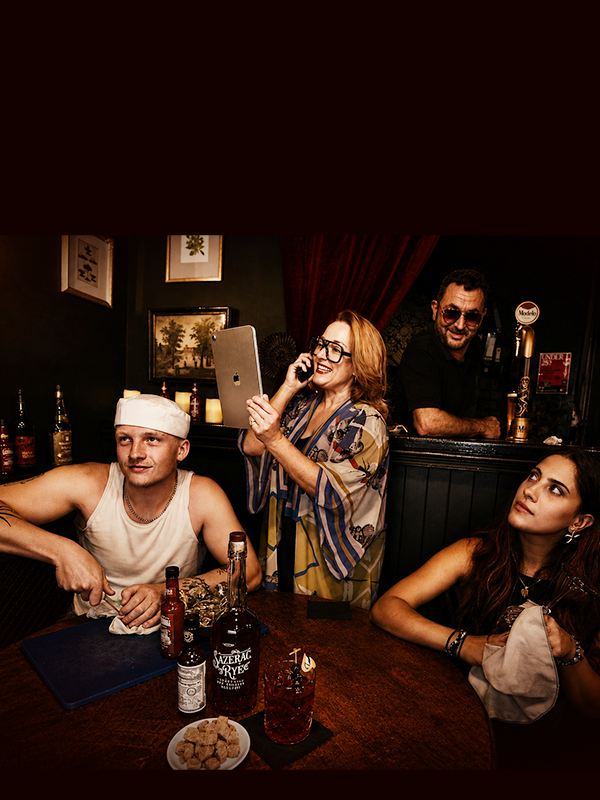 Group of four people in a bar, one man with a white chef's hat, a woman holding a phone, a woman with long dark hair, and a bartender smiling behind the bar