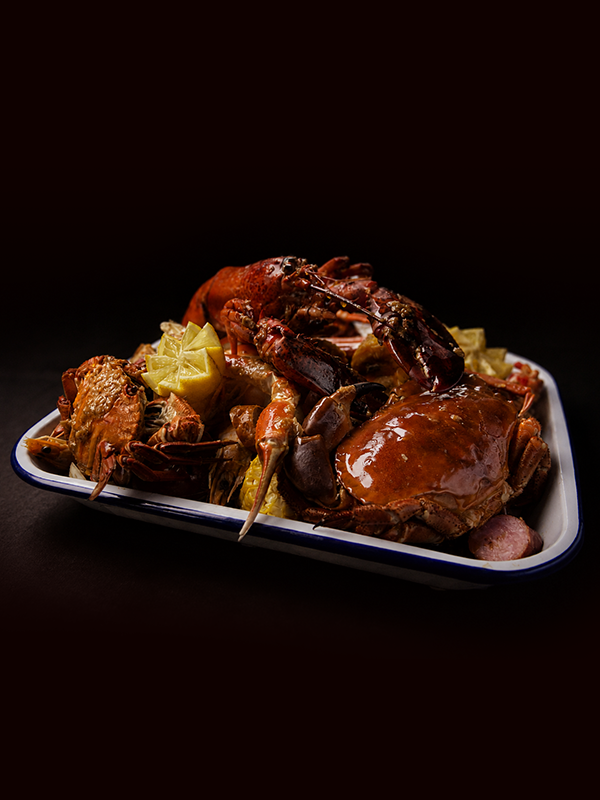 A seafood platter featuring a crab with a glossy sauce, a lobster tail, and lemon wedges, served on a white plate against a dark background.
