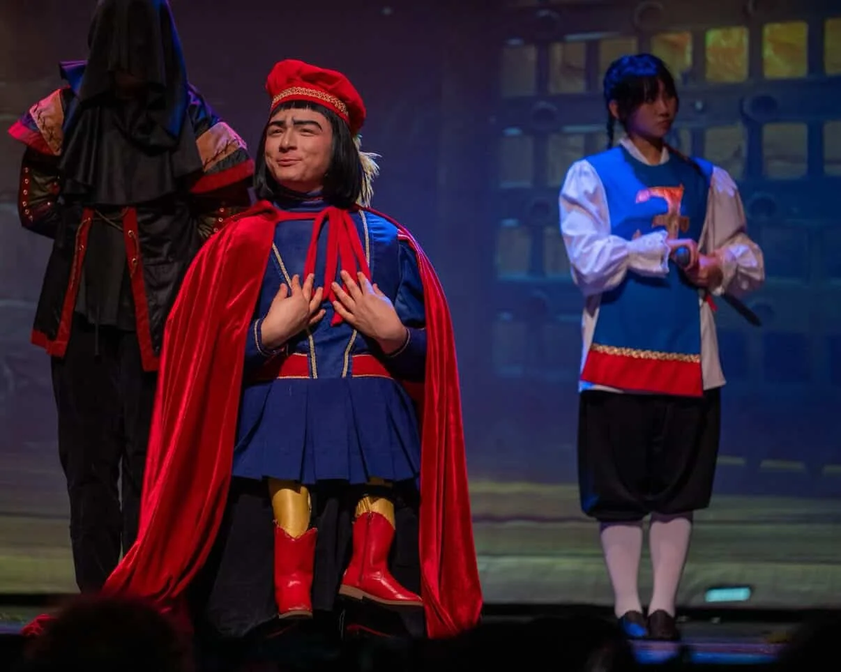 Children dressed in theatrical costumes perform on stage, with one wearing a blue and red outfit and a red cape, expressing a dramatic emotion.