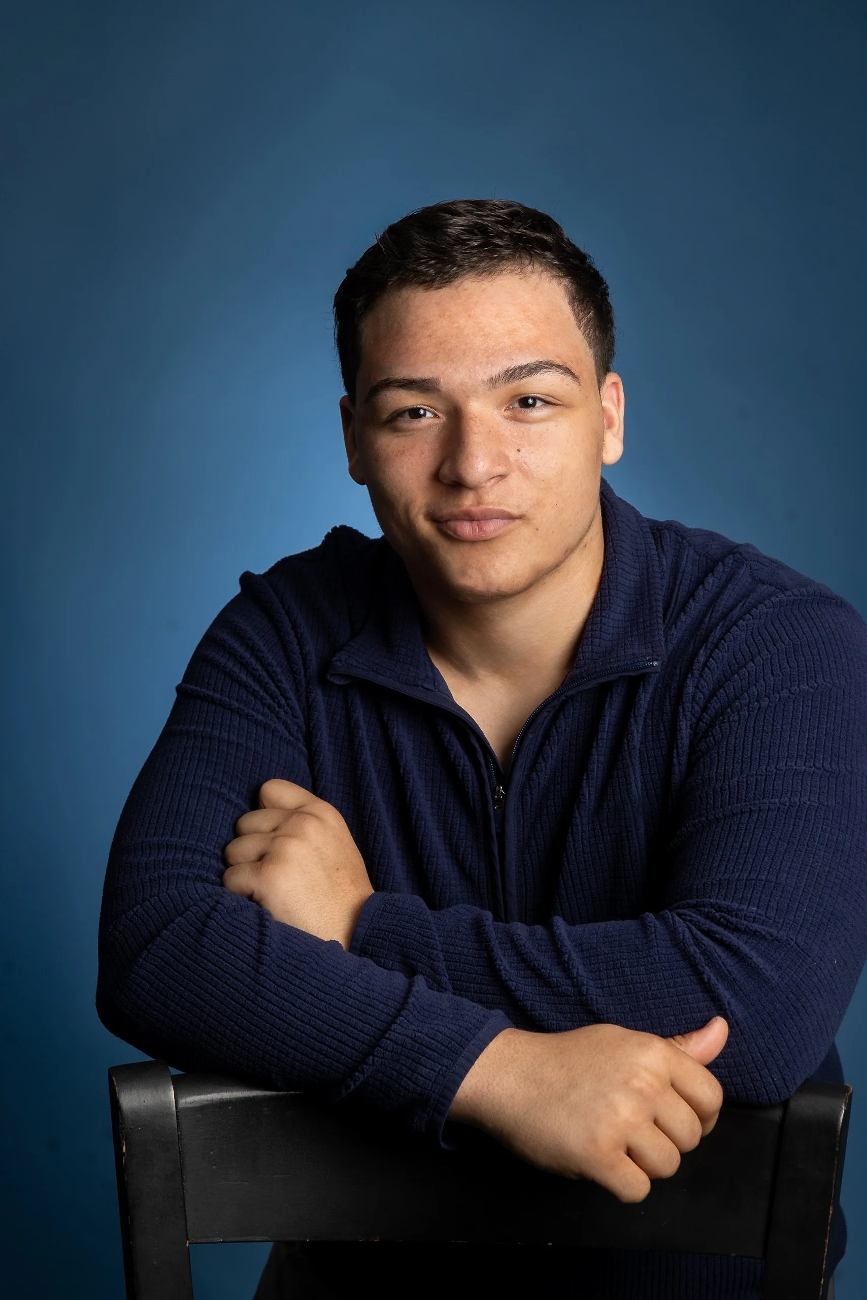 Young man with short dark hair, fair skin, and freckles, wearing a navy blue zip-up sweater, sitting in front of a dark blue background, looking directly at the camera with a slight smile, crossing his arms on a black chair.