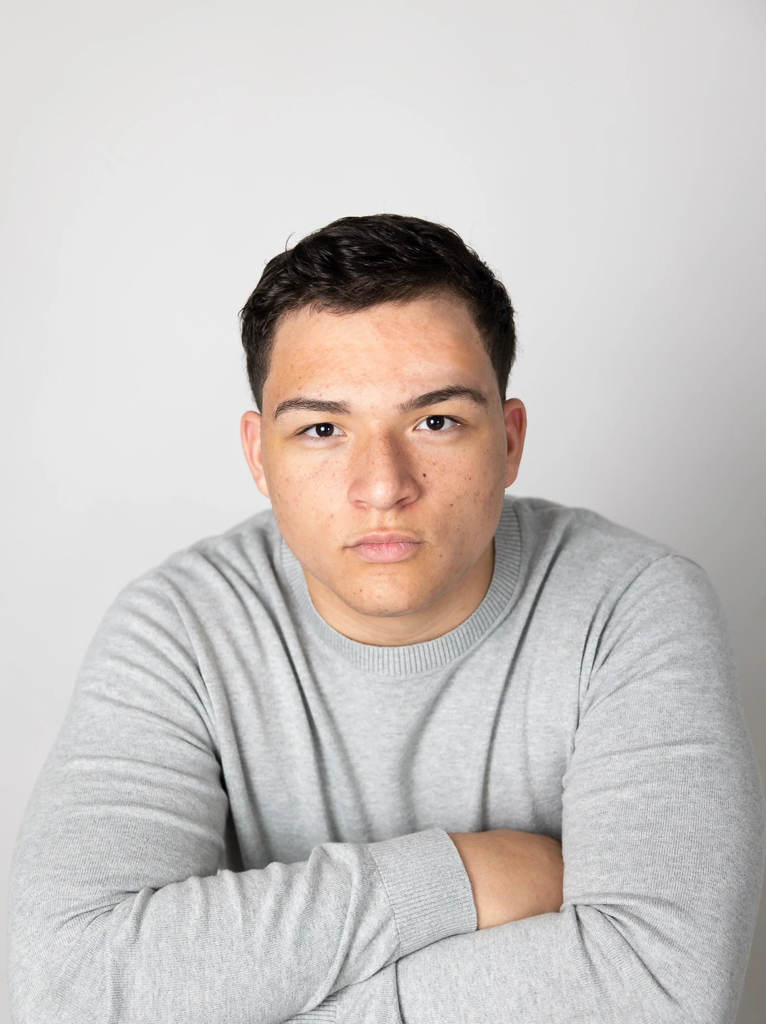 A young man with short dark hair and freckles, wearing a gray sweater, sitting with crossed arms against a plain white background.