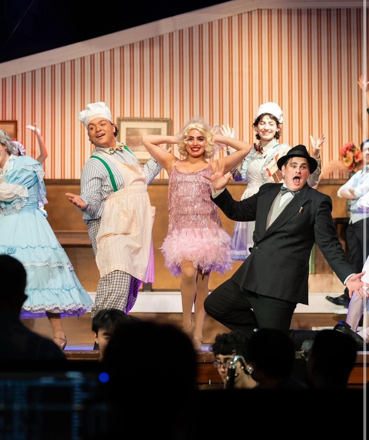 The image shows actors performing on stage in a theatrical musical, with three main characters in colorful costumes and expressive poses, and a backdrop of a striped wall.