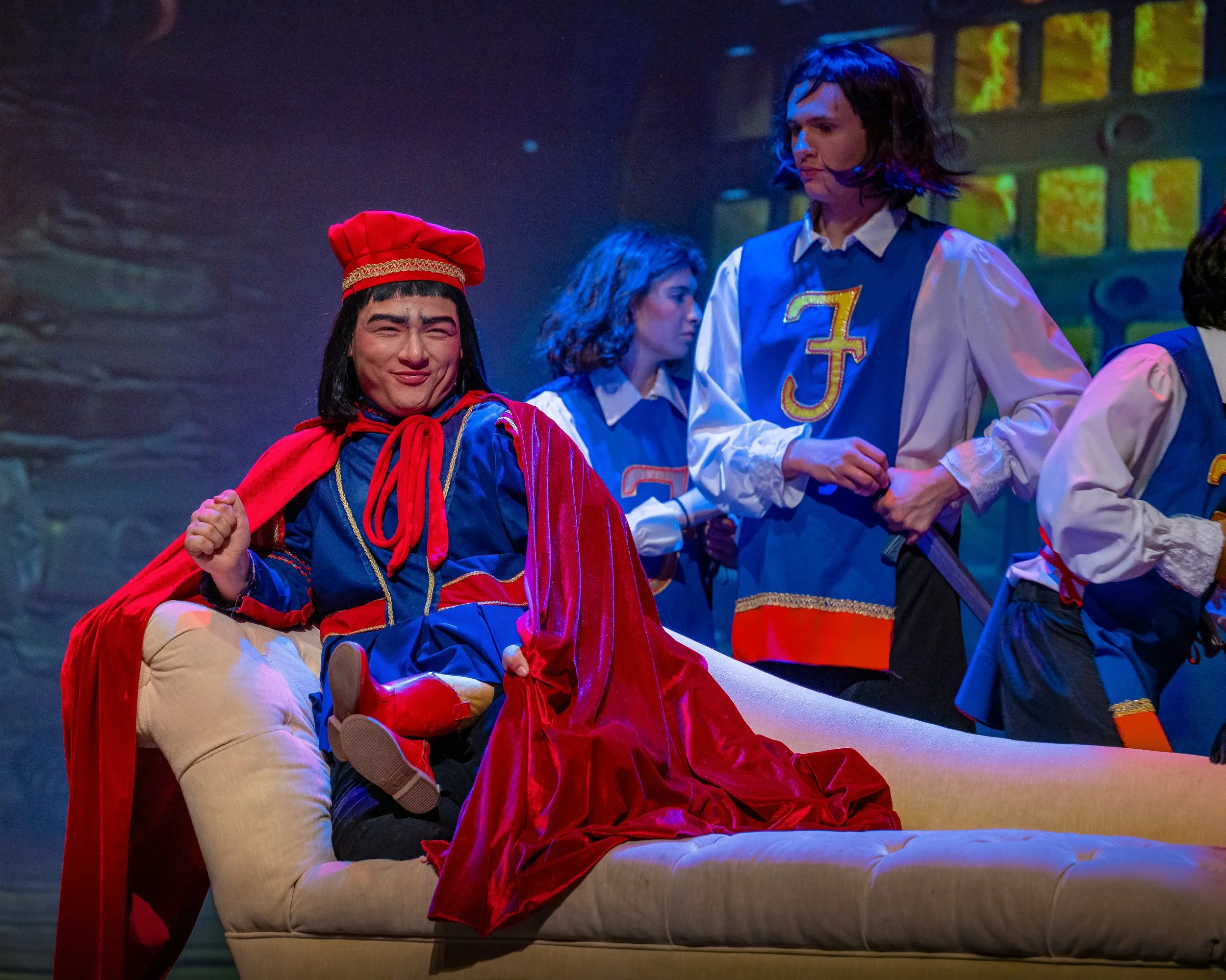 Scene from a theatrical play with actors dressed in costumes, including one person in a red cape and hat, and others in blue and white outfits, on stage with a colorful background.