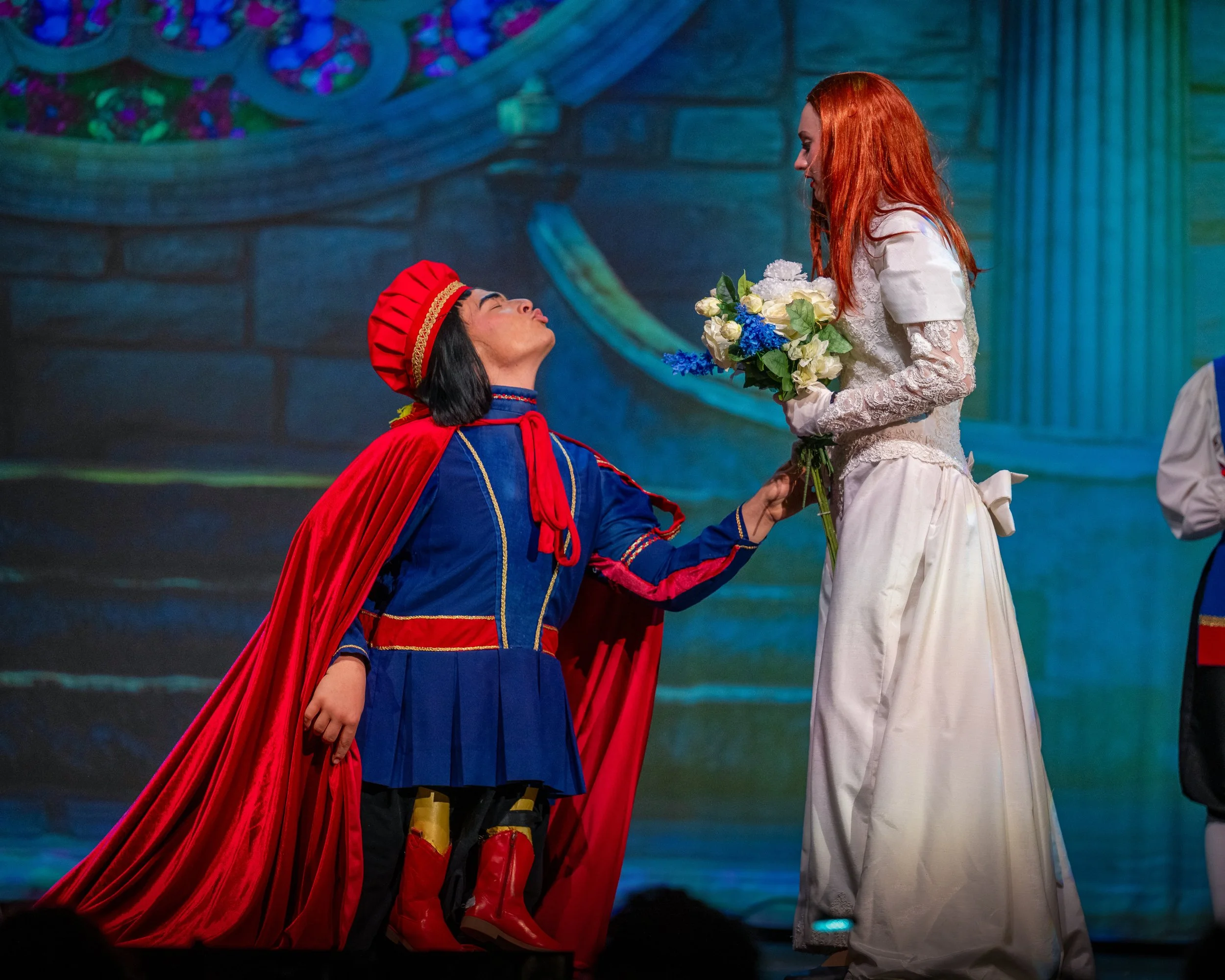A scene from a theatrical performance showing a person dressed in a colorful costume kneeling and kneeling, and holding the hand of a woman with red hair, who is wearing a white lace dress and holding a bouquet of white and blue flowers. The backgrou