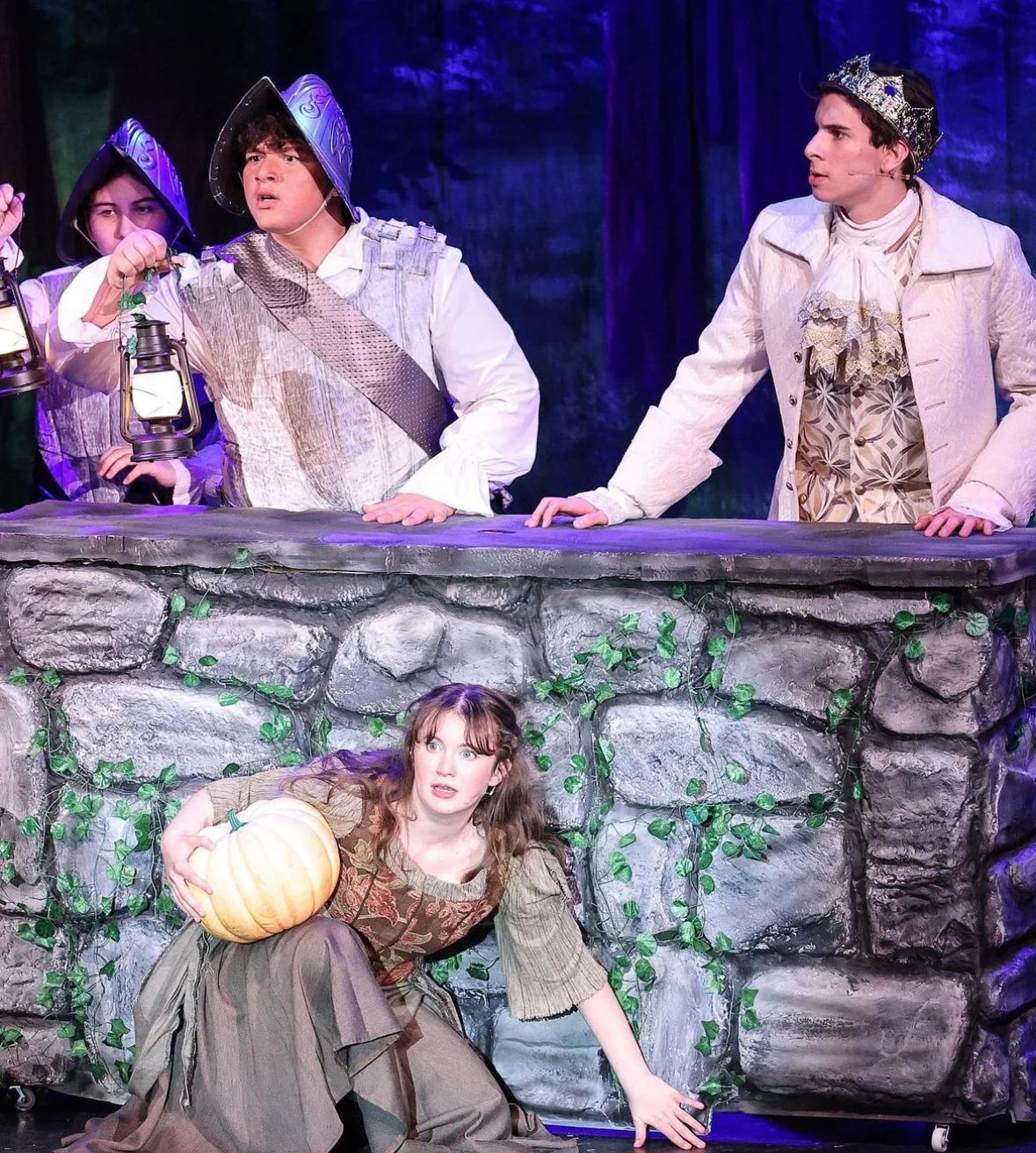 The image depicts a theatrical scene with four actors on stage. Three actors are standing behind a stone-like set piece, with one of them wearing a crown and costumes resembling royalty, holding a lantern. The other two actors also wear period costum