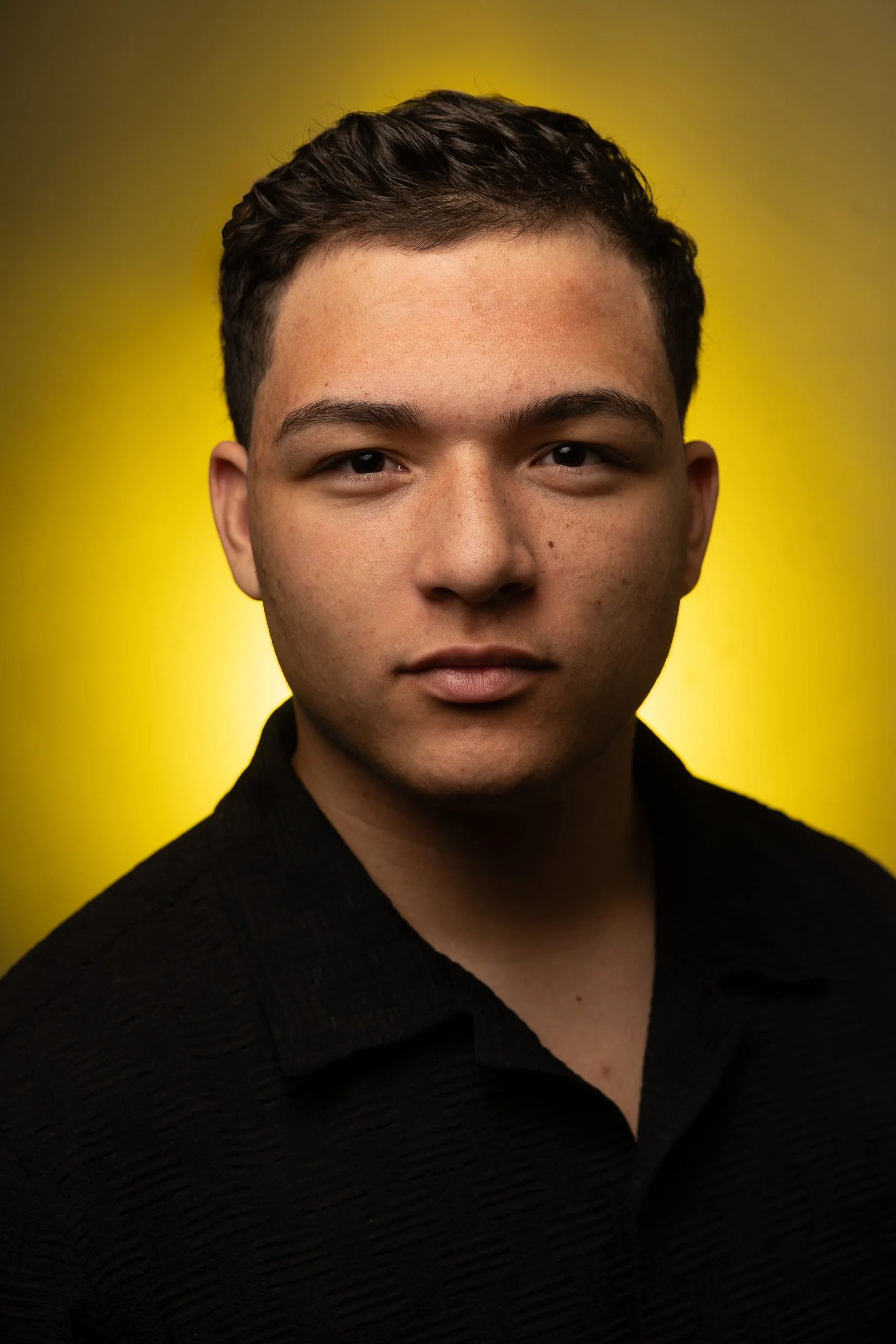 Close-up portrait of a young man with short dark hair, wearing a black textured shirt, set against a gradient yellow background.