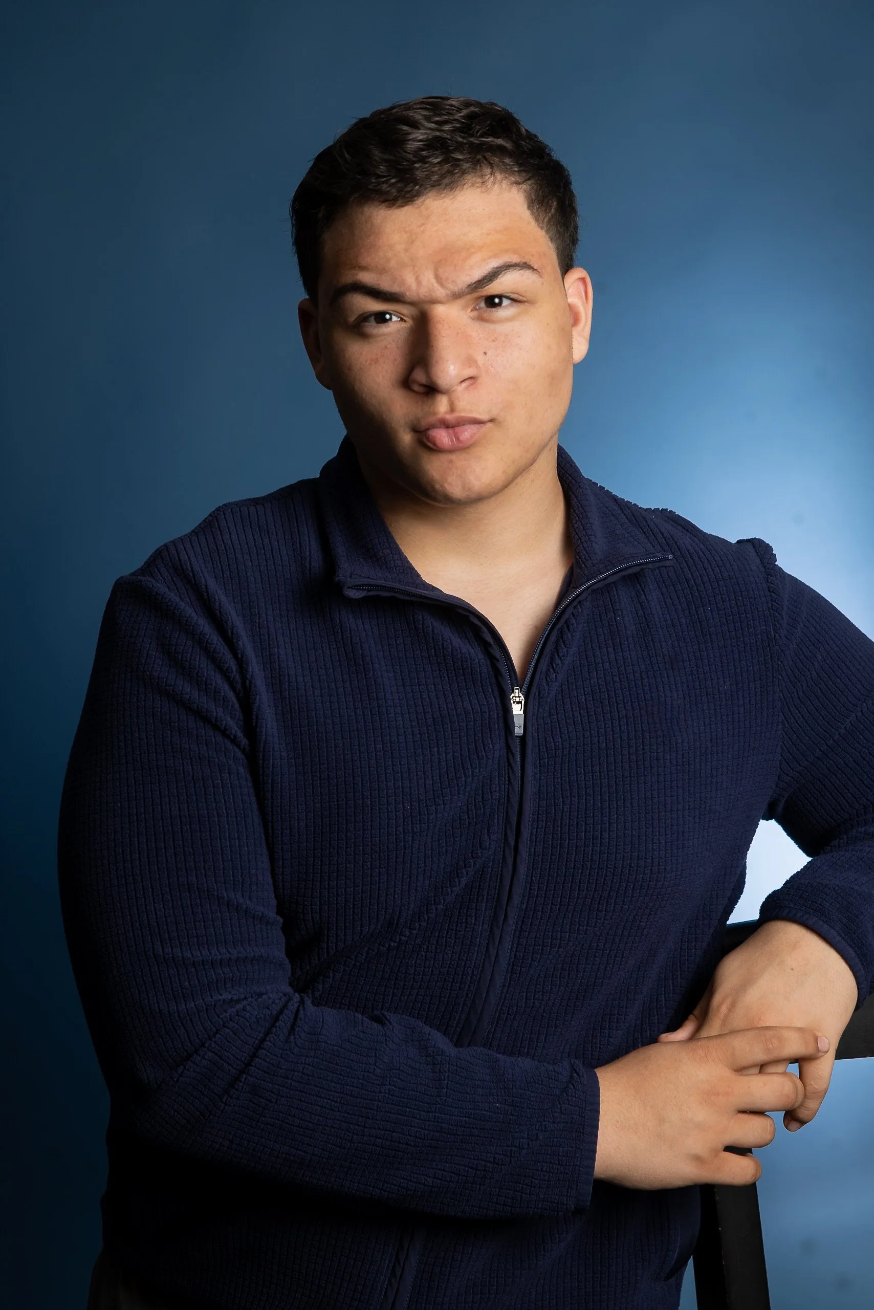 A young man with short dark hair making a duck face, wearing a navy blue zip-up sweater, against a blue background.