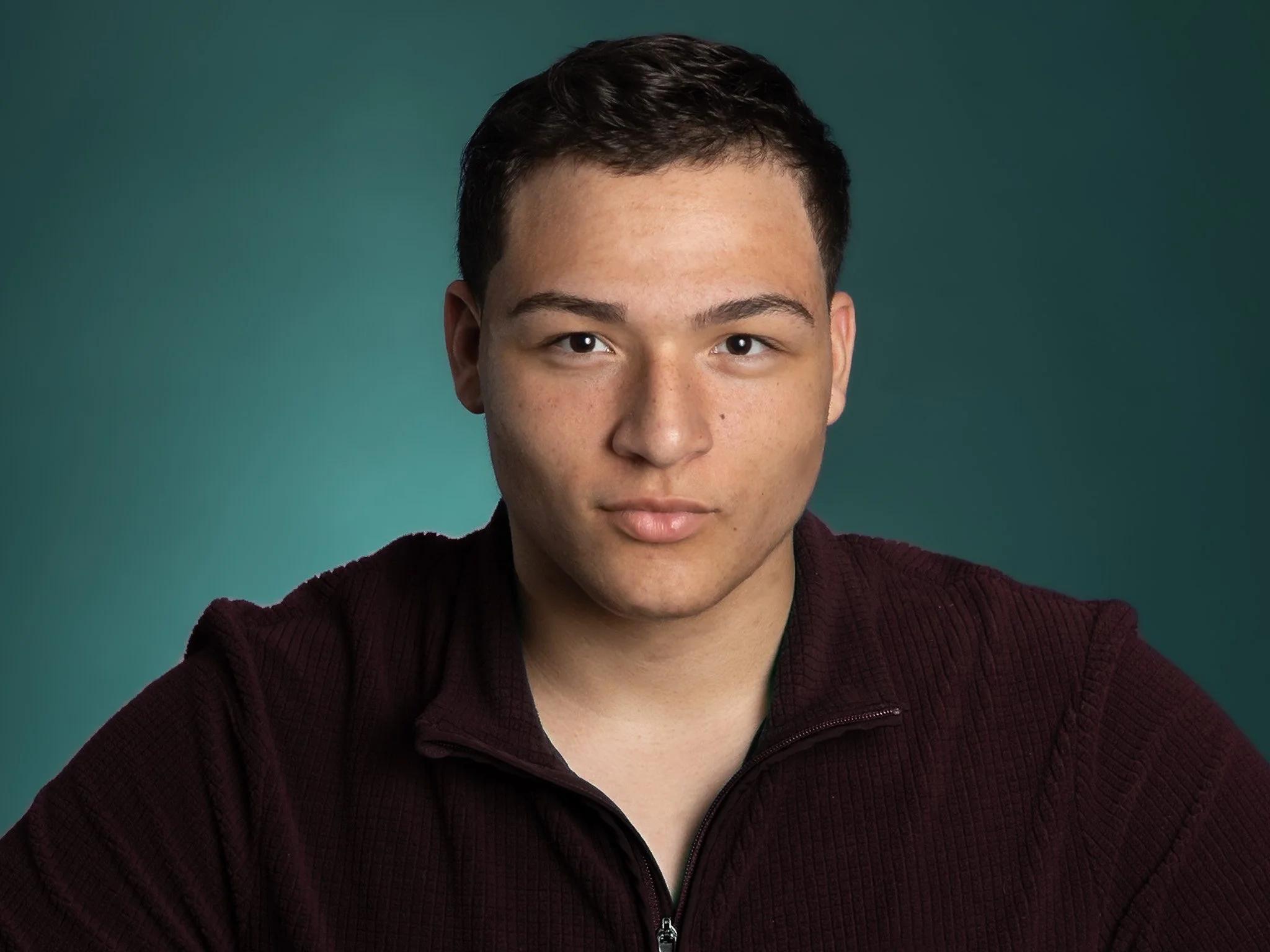 Close-up of a young man with short dark hair and light skin, wearing a maroon zip-up sweater, looking directly at the camera against a teal background.