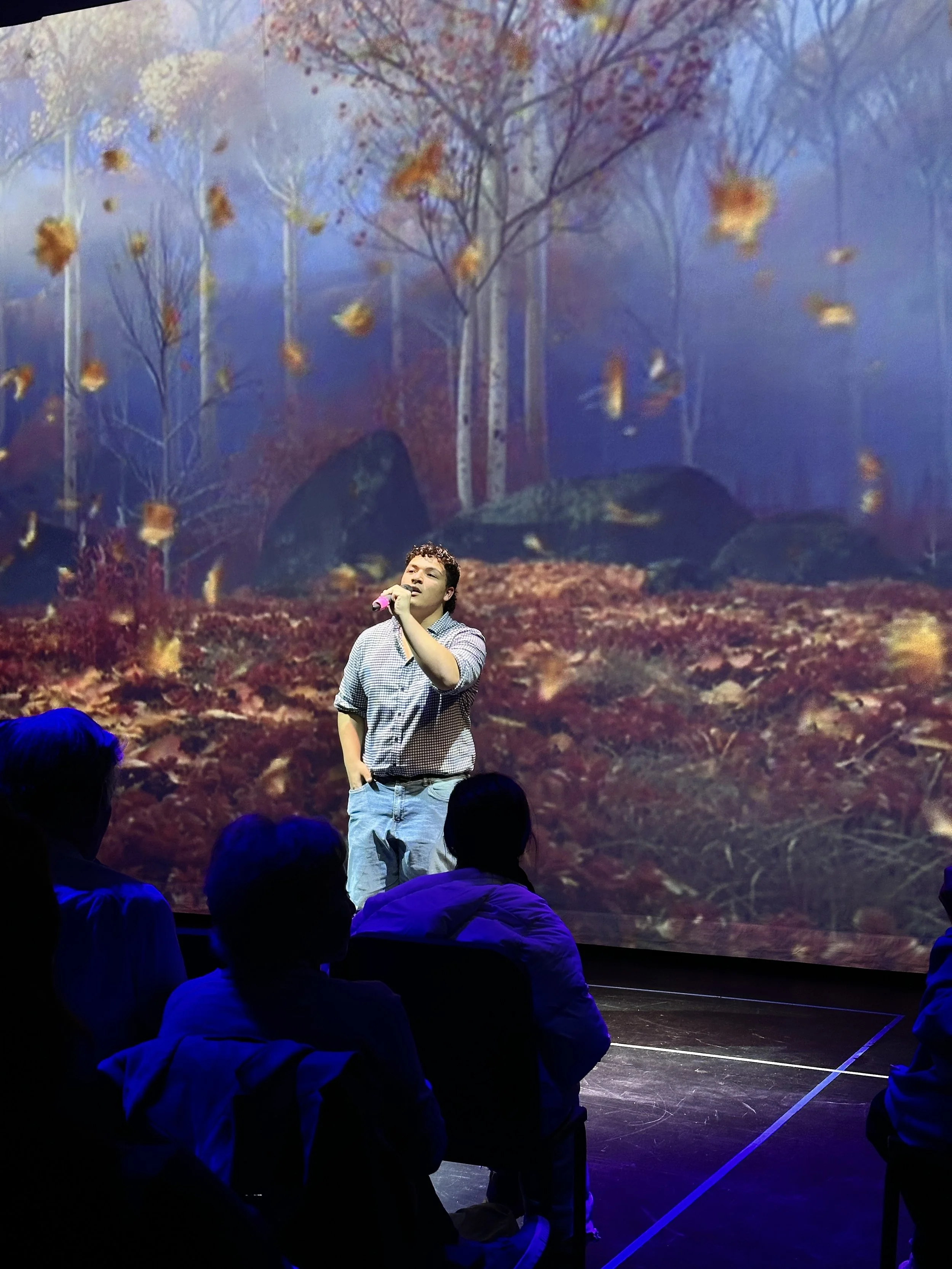 A man singing into a microphone on stage, with a background of a forest scene featuring trees and falling leaves.