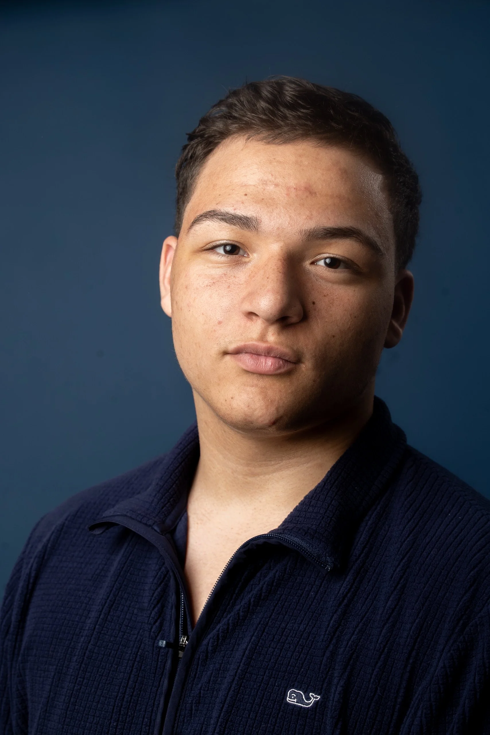 Close-up of a young man with short dark hair and light skin, wearing a navy blue zip-up jacket with a small white whale logo, standing against a dark blue background.